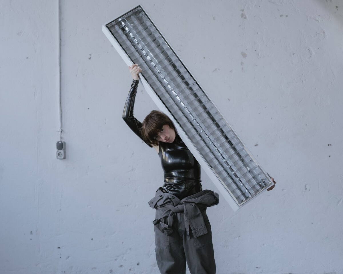 Eine Person in schwarzem Latexoberteil und grauer Arbeitshose steht vor einer hellen Wand und hält ein langes, rechteckiges Deckenleuchtpanel schräg über der Schulter.