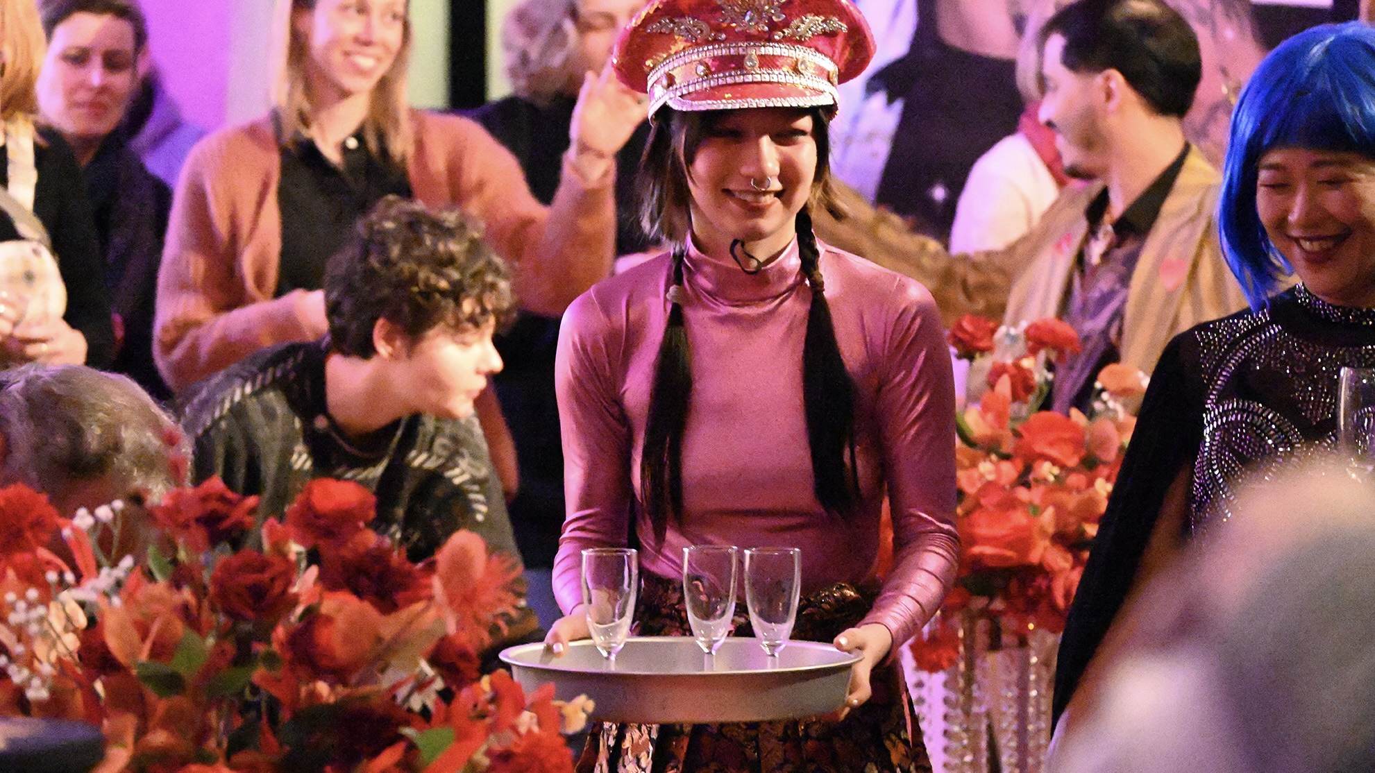 Eine Frau in glänzendem violetten Oberteil und rotem Kopfschmuck trägt ein Tablett mit Gläsern durch einen festlich dekorierten Raum voller Menschen und roter Blumen.
