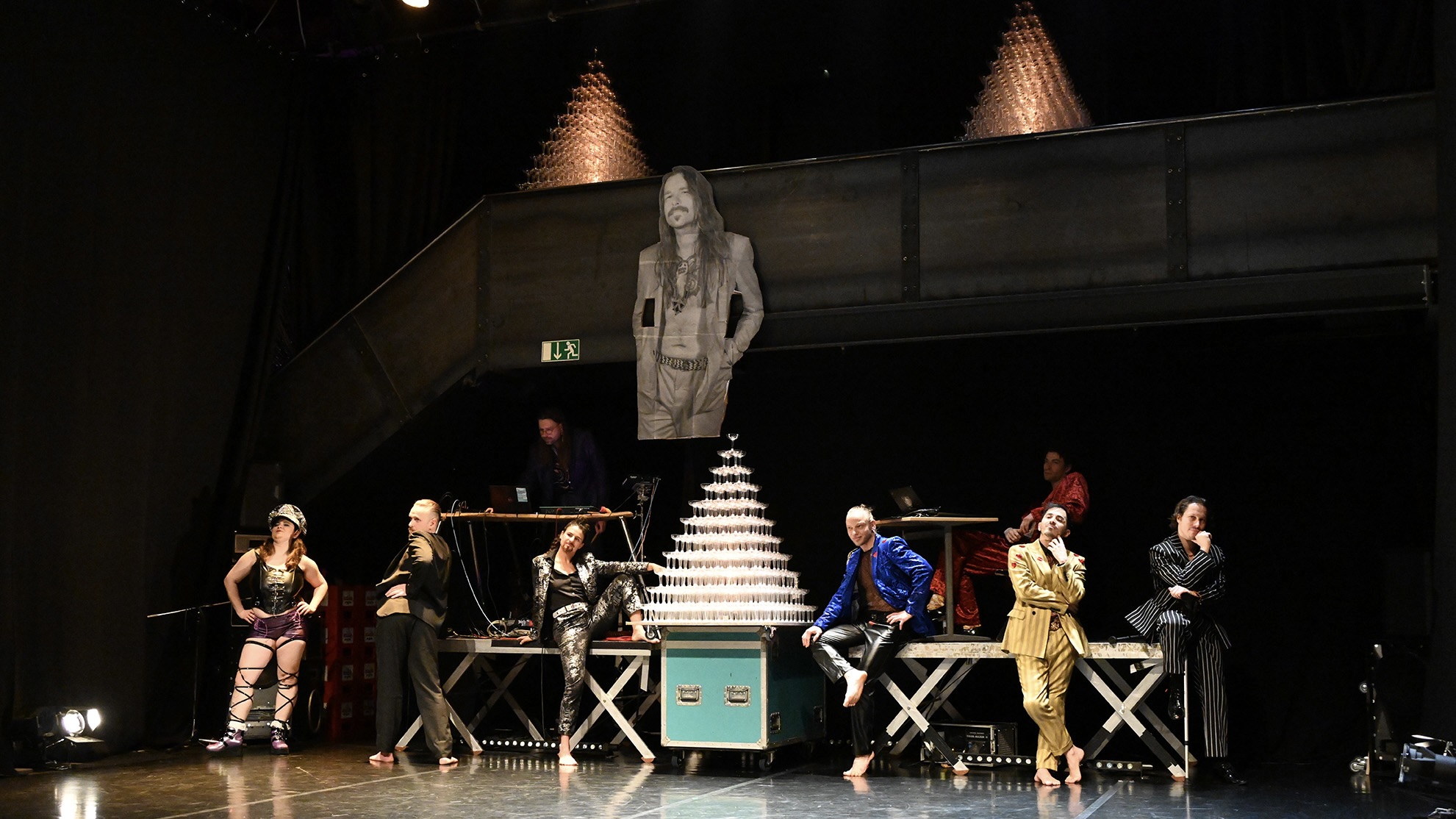Mehrere Darsteller*innen in auffälligen Kostümen stehen, sitzen und posieren vor einem großen Turm aus Sektgläsern auf einer Theaterbühne; im Hintergrund ist eine Galerie mit Lichtinstallation zu sehen.