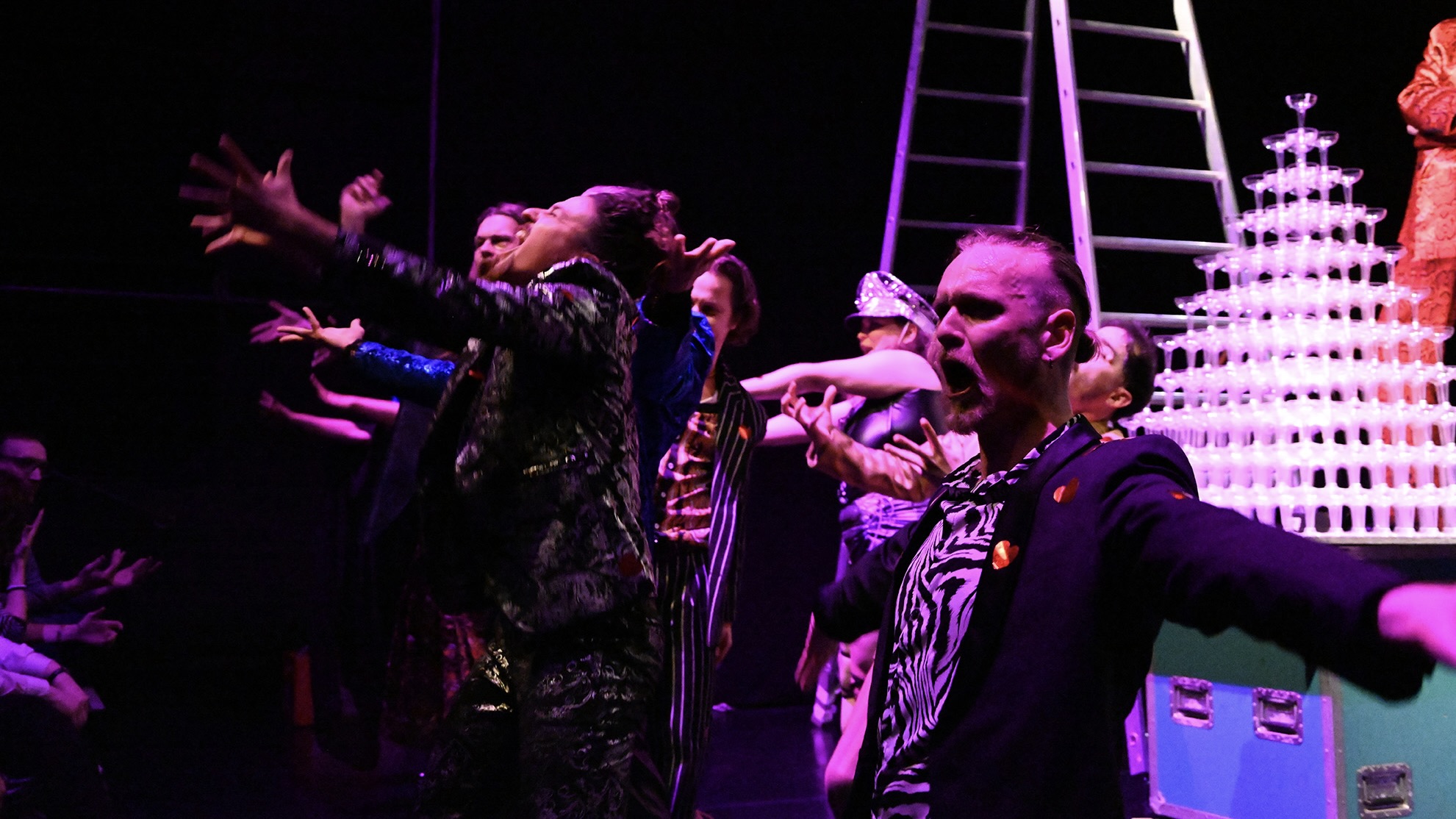 Eine Gruppe von Darsteller*innen in farbenfrohen Kostümen streckt jubelnd die Arme nach vorne. Im Hintergrund sind ein Gläserturm und eine Leiter auf der Bühne sichtbar.