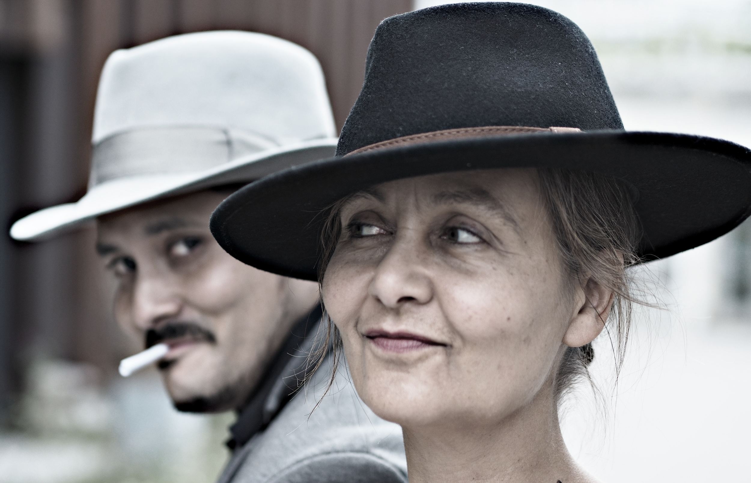 Ein Mann und eine Frau mit unterschiedlichen, breitrandigen Hüten stehen nah beieinander, wobei nur ein Teil der Gesichter zu sehen ist. Die Bildgestaltung setzt auf helle und dunkle Kontraste, der Hintergrund bleibt unscharf. Der Mann lächelt im Hintergrund; er hat eine Zigarette im Mund.