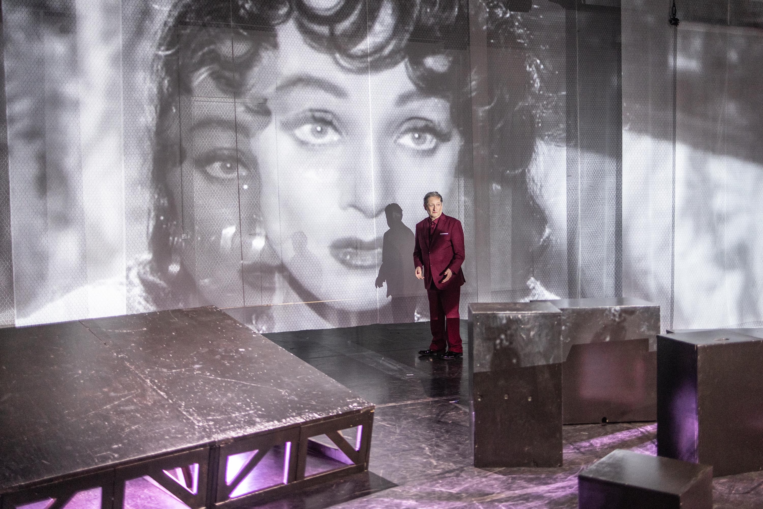 Eine Theaterbühne mit schwarzen Podesten vor einer groß projizierten schwarz-weißen Filmaufnahme eines Gesichts, in der Bildmitte verdeckt ein graues Rechteck einen Teil der Projektion. Eine Person im roten Anzug steht vor der Leinwand