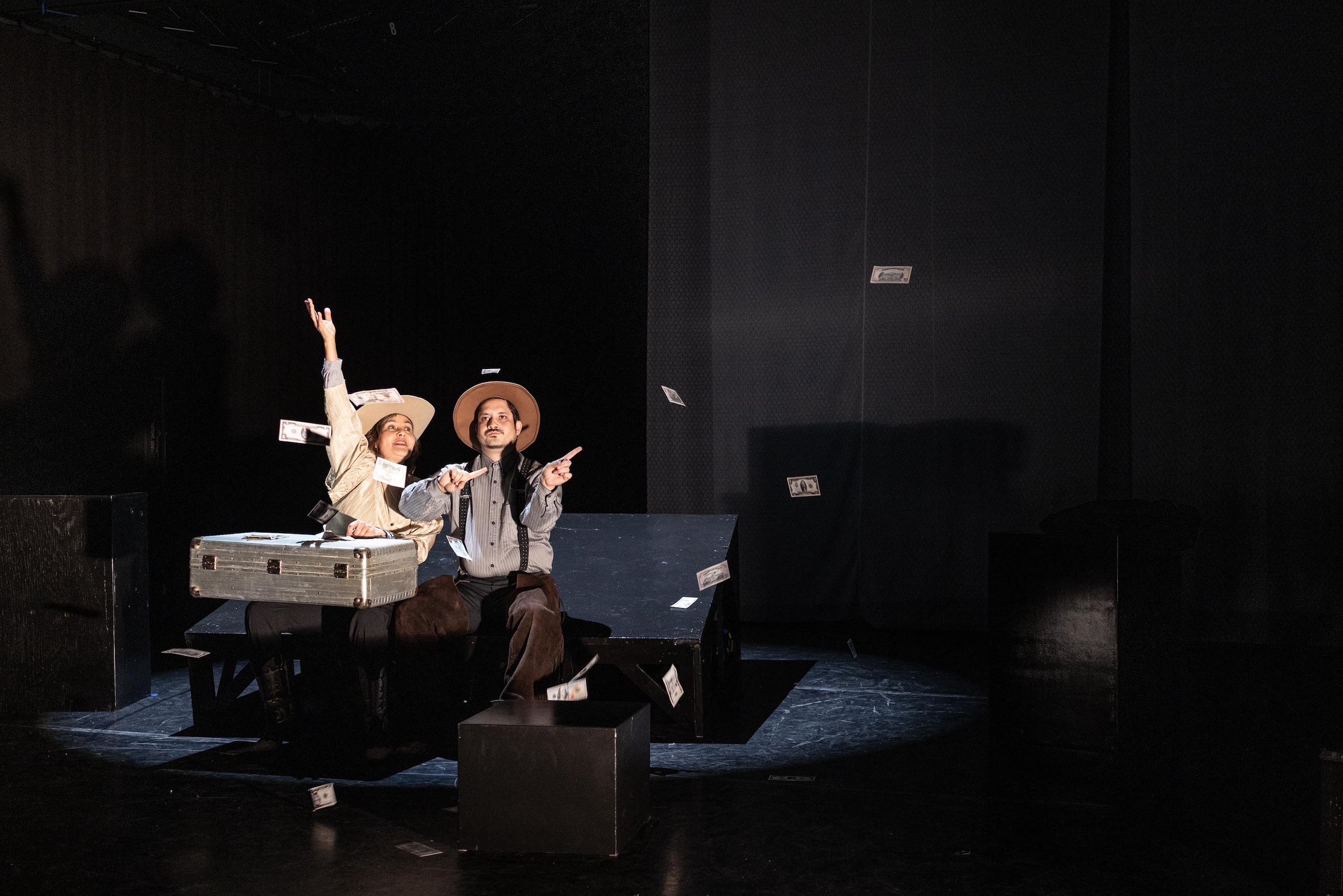Zwei Personen in Anzügen und Hüten sitzen auf einer Theaterbühne, umgeben von schwebenden Geldscheinen, vor ihnen ein geöffneter silberner Koffer, die Szene ist dramatisch ausgeleuchtet.