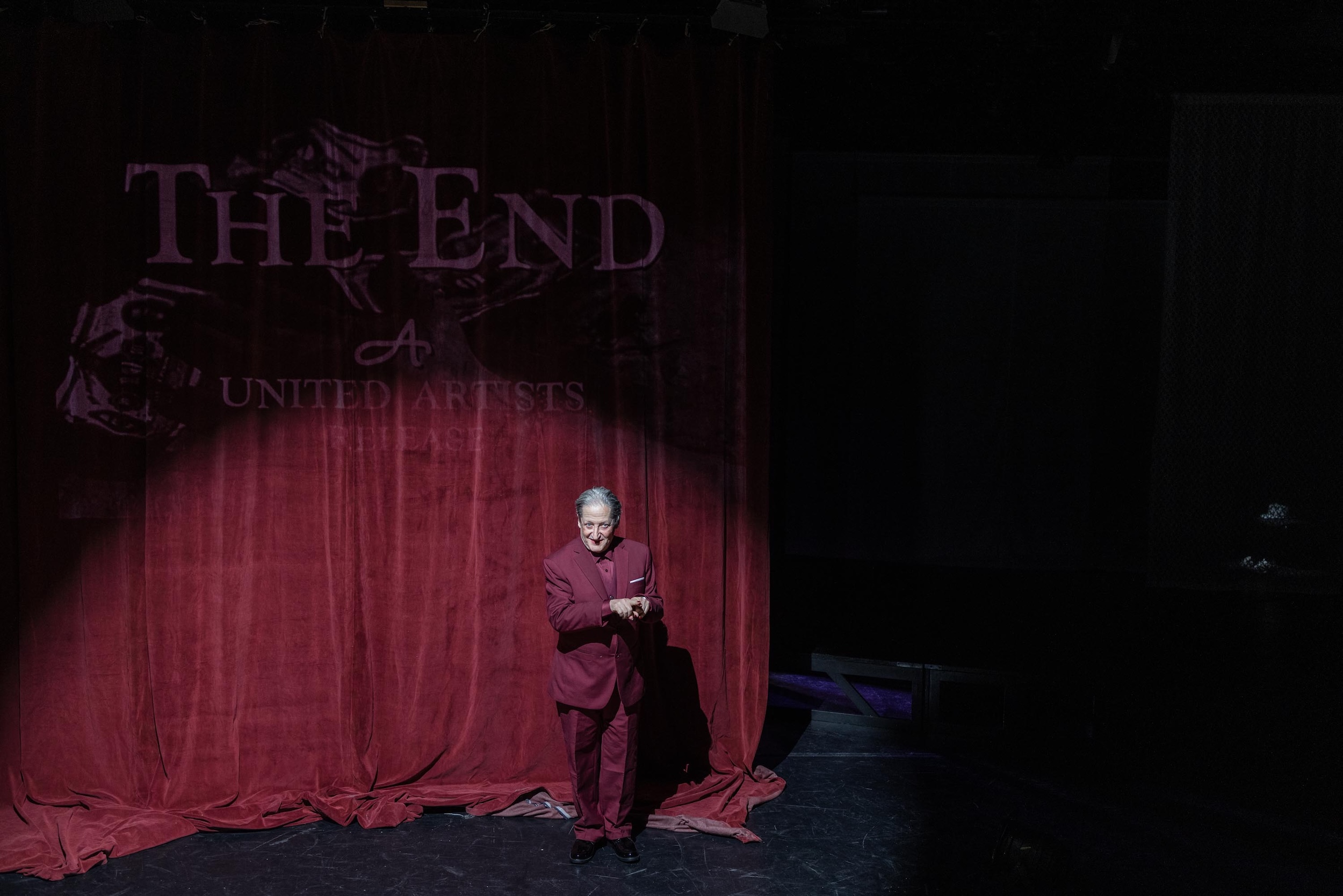Eine Person in dunklem Anzug steht allein im Lichtkegel vor einem schweren roten Vorhang mit der groß projizierten Aufschrift „THE END – A UNITED ARTISTS RELEASE“.