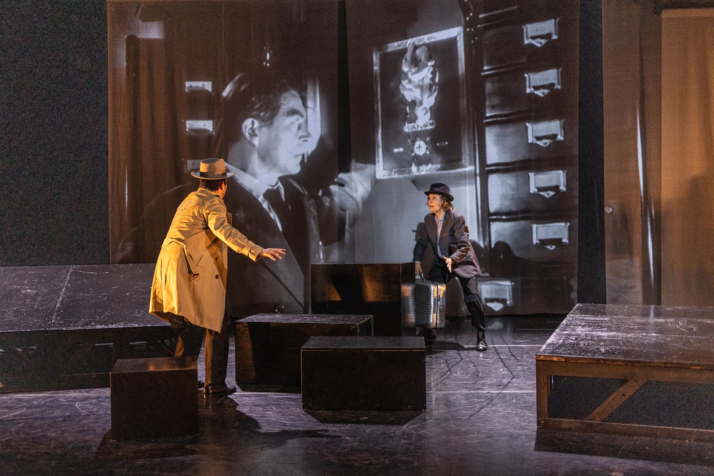 Zwei Personen in Mänteln und Hüten agieren auf einer Theaterbühne mit Podesten und silbernem Koffer, im Hintergrund läuft eine schwarz-weiße Filmprojektion, die teilweise von einem grauen Rechteck überdeckt wird.