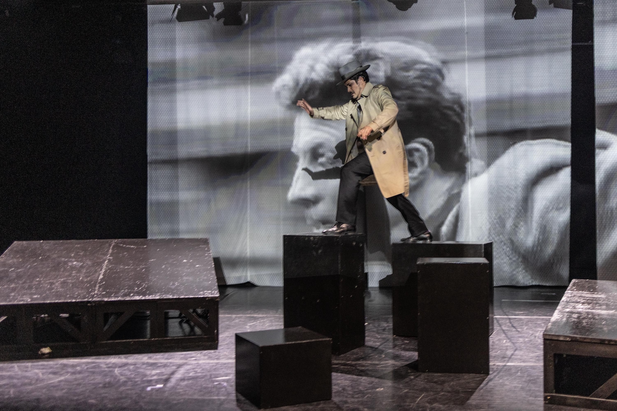 Eine Person im langen Mantel und mit Hut balanciert auf schwarzen Podesten einer Theaterbühne, im Hintergrund ist ein groß projiziertes schwarz-weißes Filmgesicht zu sehen.