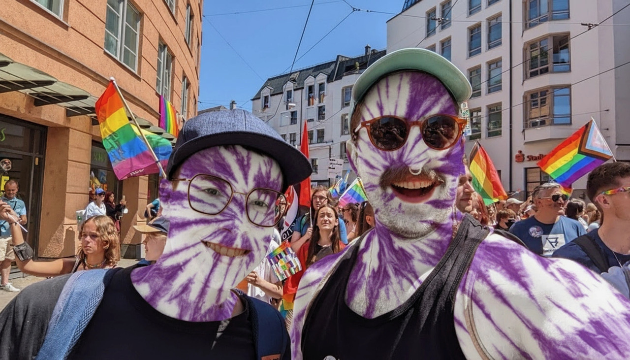 Zwei Personen bei einer bunten Pride-Parade, ihre Gesichter und Körper sind auffällig lila-weiß bemalt. Beide tragen Sonnenbrillen, eine Person außerdem einen blauen Hut. Im Hintergrund sind viele weitere Teilnehmende sowie Regenbogenfahnen und Progress-Flags zu sehen, städtische Häuser und eine sonnige Straße bilden das Umfeld. 