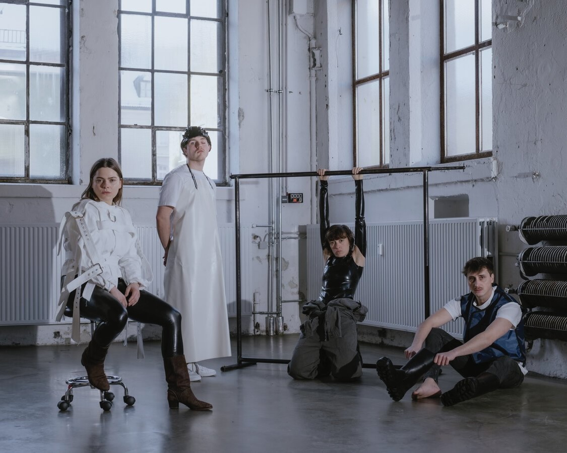 Vier Personen posieren in einem hohen, industriellen Raum mit großen Fenstern; eine sitzt in Zwangsjacke auf einem Hocker, eine steht in weißer Schürze, eine hängt an einer Metallstange, eine sitzt am Boden in Gummistiefeln.