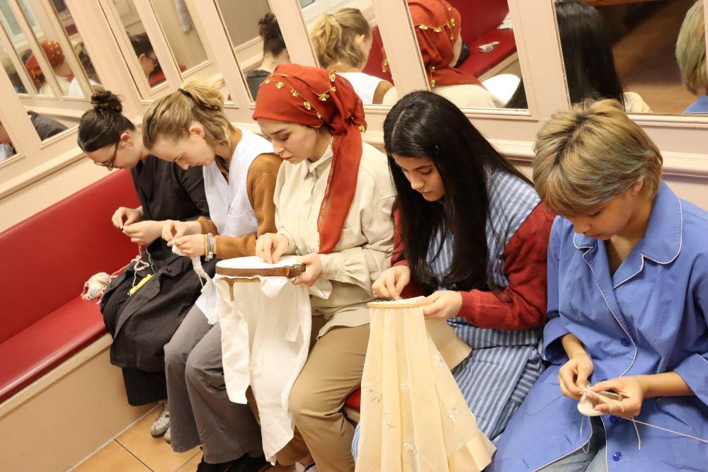 Fünf Personen sitzen nebeneinander auf einer roten Bank und sticken oder nähen gemeinsam, im Hintergrund Spiegel an der Wand