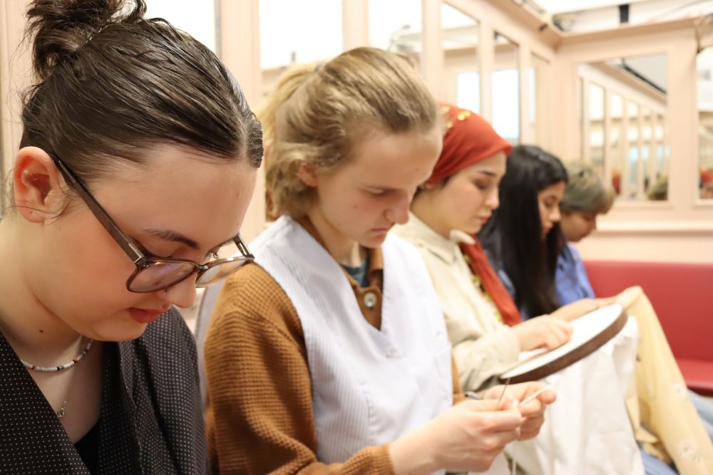 Nahaufnahme von vier Personen, die konzentriert auf einer Bank sitzen und sticken, ein Teil von ihnen trägt Arbeitskittel oder Kopftuch