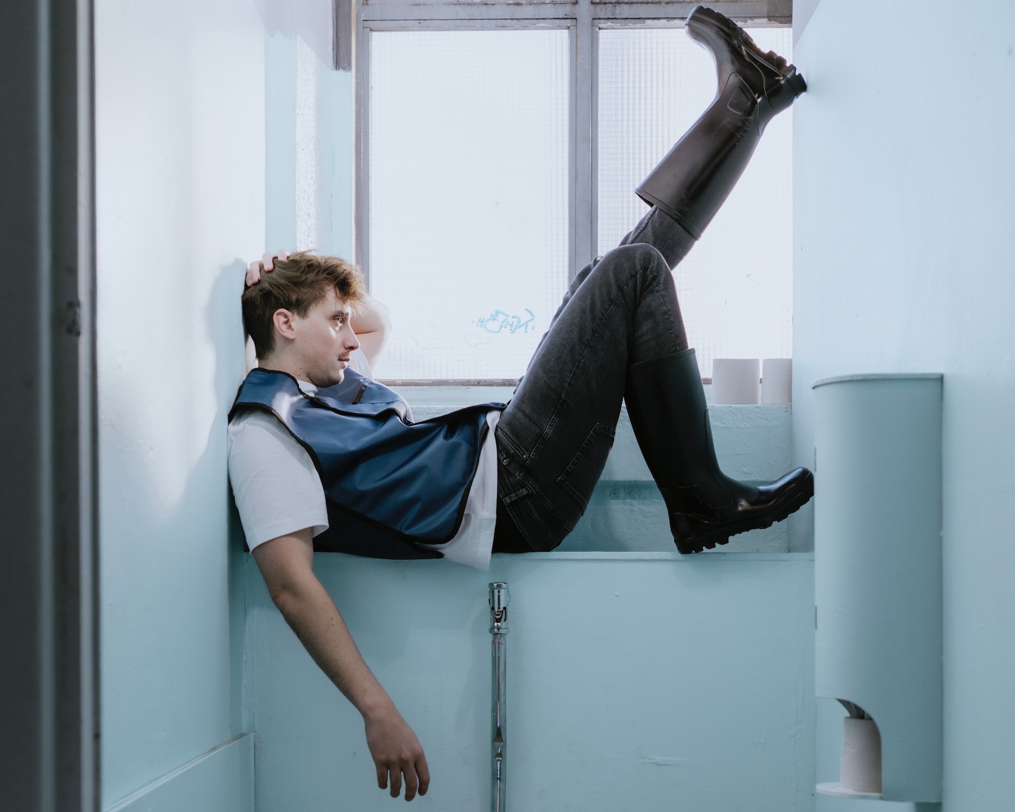 Eine Person mit kurzhaarigem, hellbraunem Haar liegt quer in einem engen, hellblauen Waschraum, stützt den Rücken an der Wand, die Füße in schwarzen Gummistiefeln gegen das Fensterbrett.