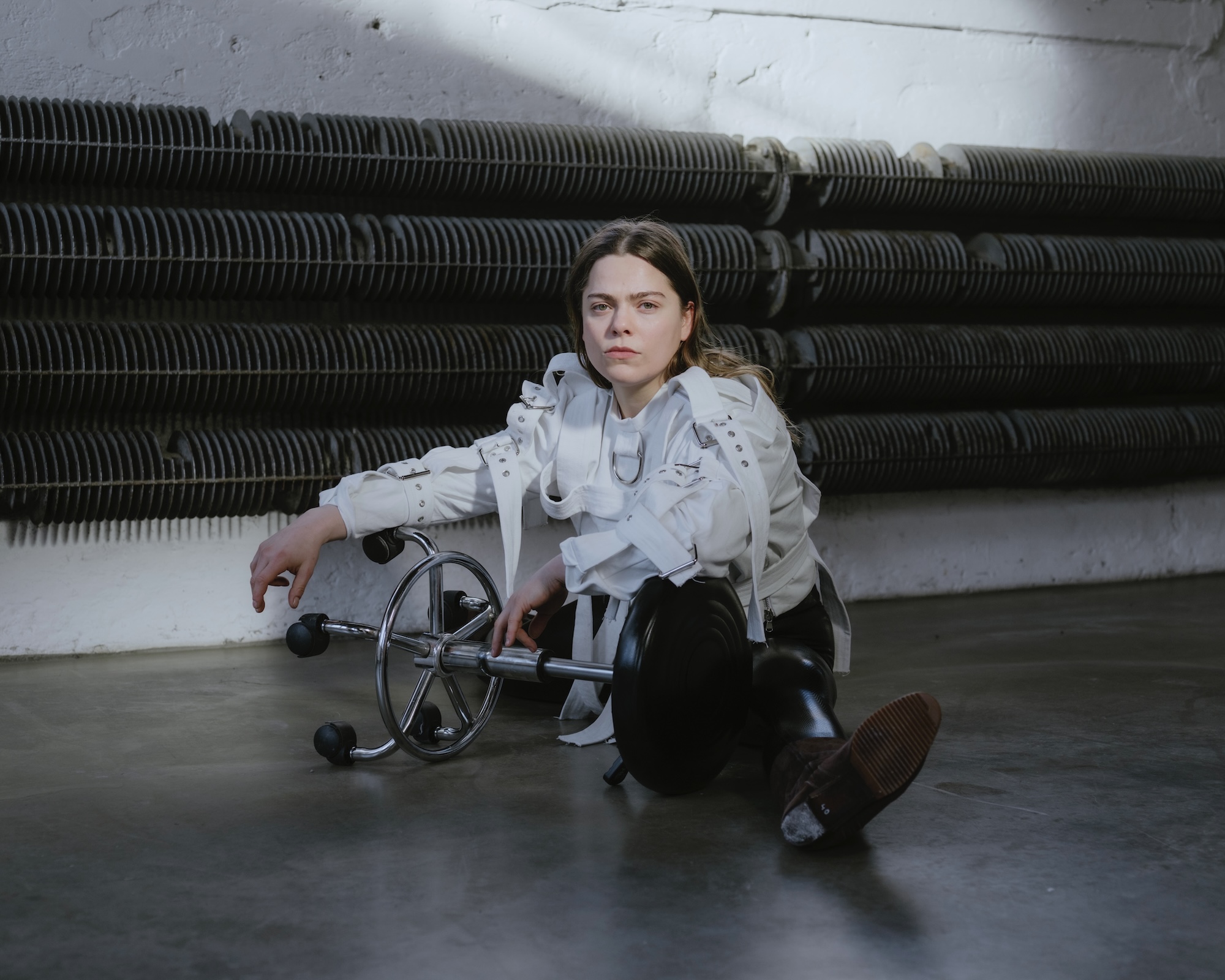 Eine Person in weißer Zwangsjacke und schwarzer Latexhose sitzt auf einem Rollhocker vor dunkel gerippten Heizkörpern und blickt ernst in die Kamera, ein Metallobjekt liegt vor ihr am Boden.