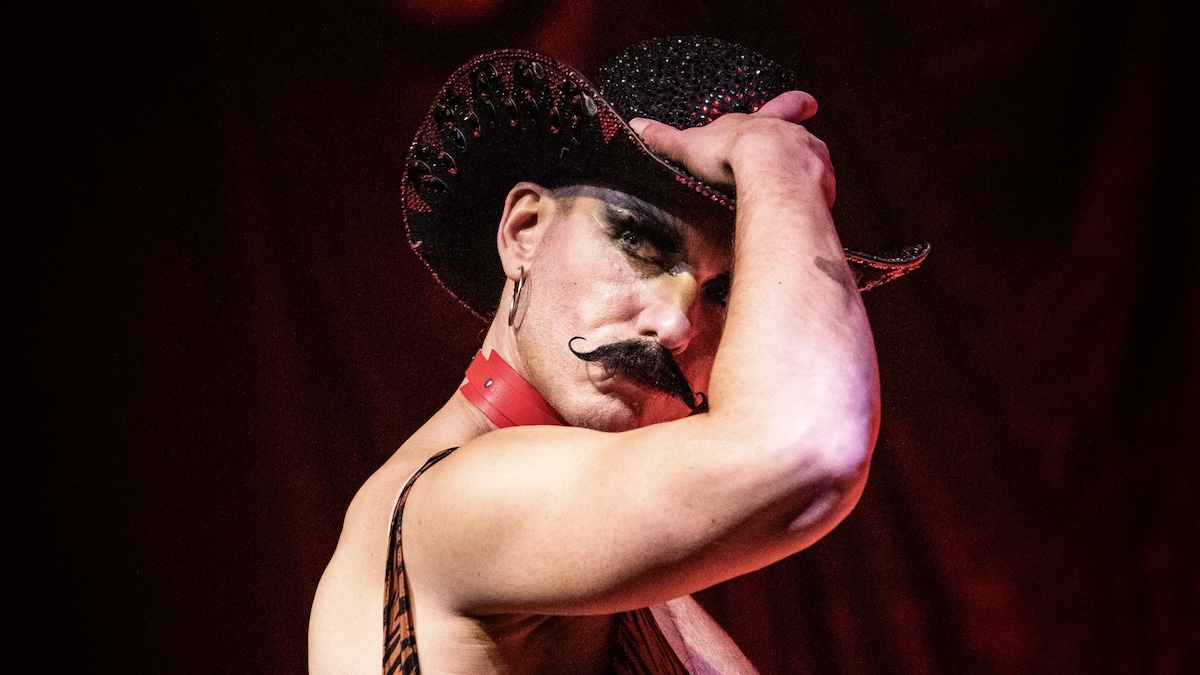 Eine Person trägt einen funkelnden, mit Strass besetzten Cowboyhut und ein rotes Halsband. Sie steht vor einem roten Hintergrund und hält den Hut mit einer Hand am Kopf fest. Zu sehen ist ein muskulöser Arm und ein Oberteil mit schmalem Träger.