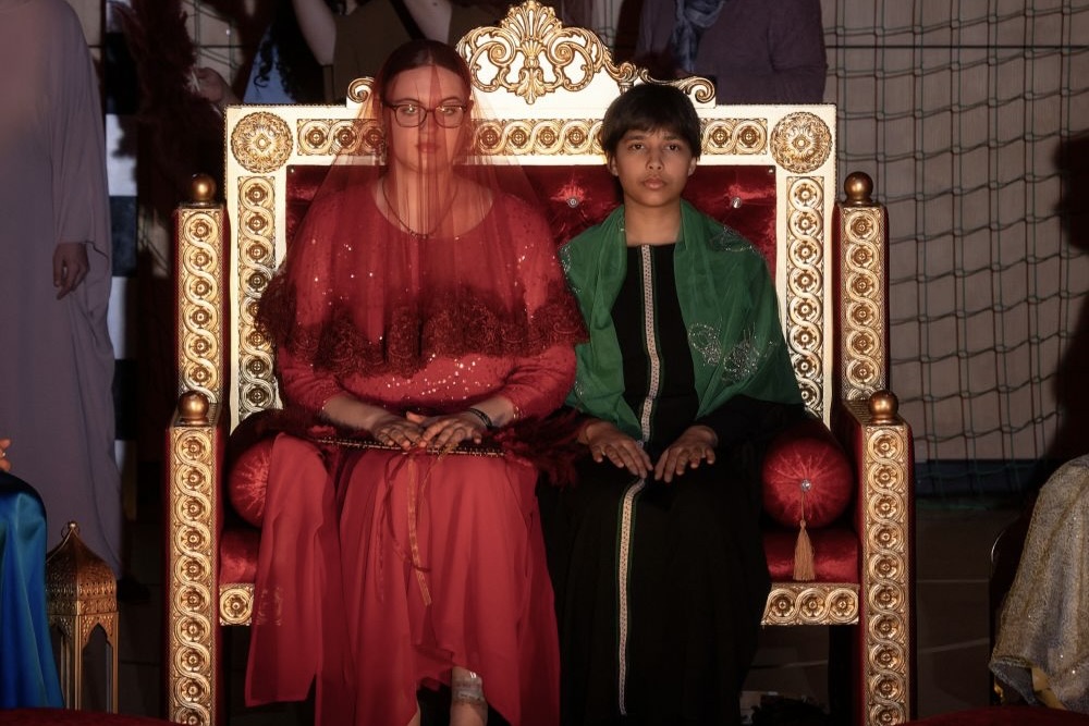 Zwei Personen in farbenprächtigen traditionellen Gewändern sitzen auf einem prunkvollen Thron, um sie herum weitere Menschen und orientalische Dekoration