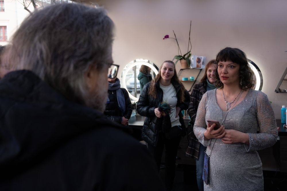 Mehrere Personen in einem hellen Raum, im Vordergrund ein Mann mit Brille, im Hintergrund eine Frau in silbernem Kleid mit Handy in der Hand, weitere Personen stehen und betrachten die Szene