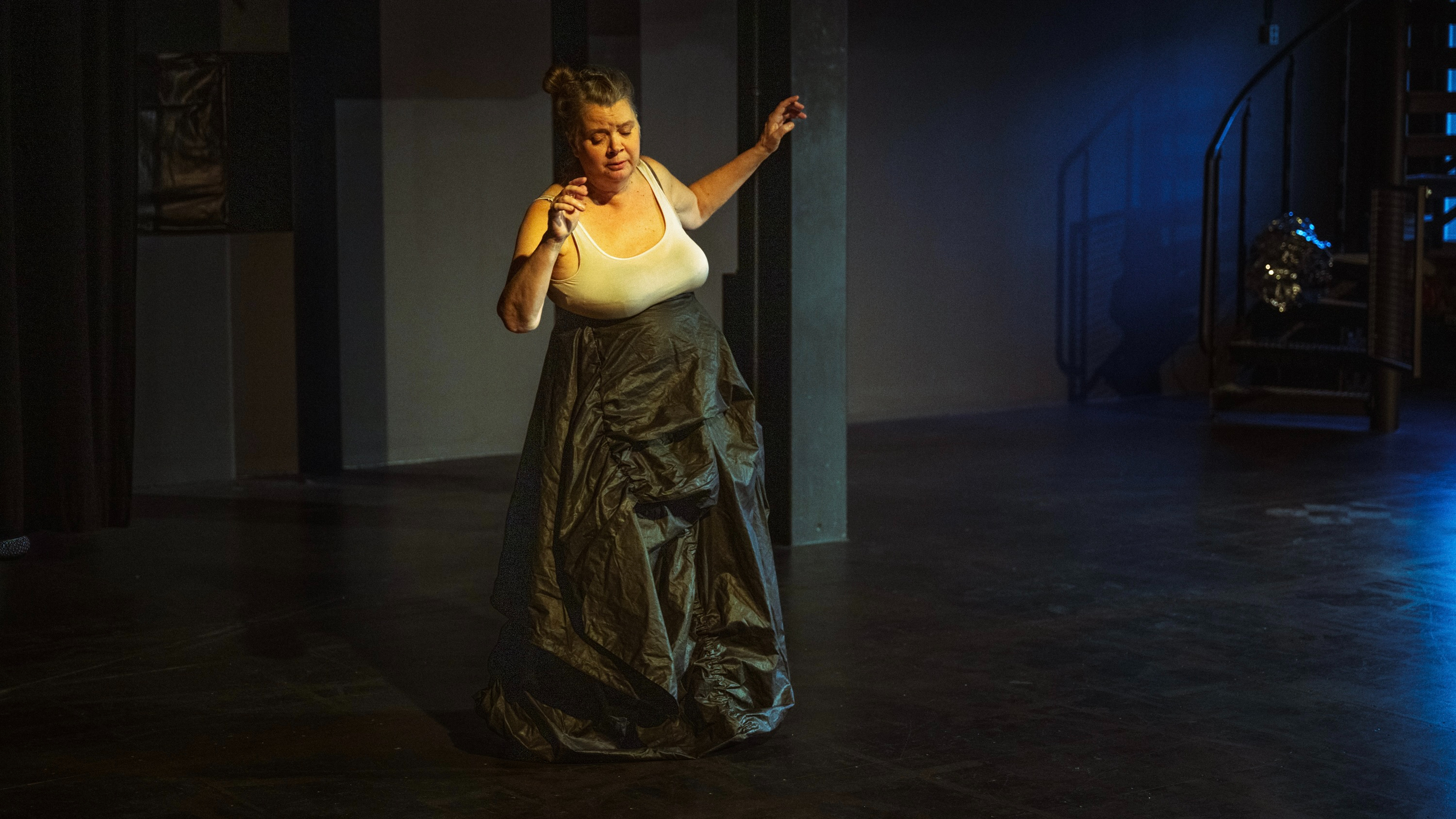 Eine Frau in einem schwarzen Rock und weißem Oberteil performt tänzerisch auf einer dunklen Bühne mit Säulen.