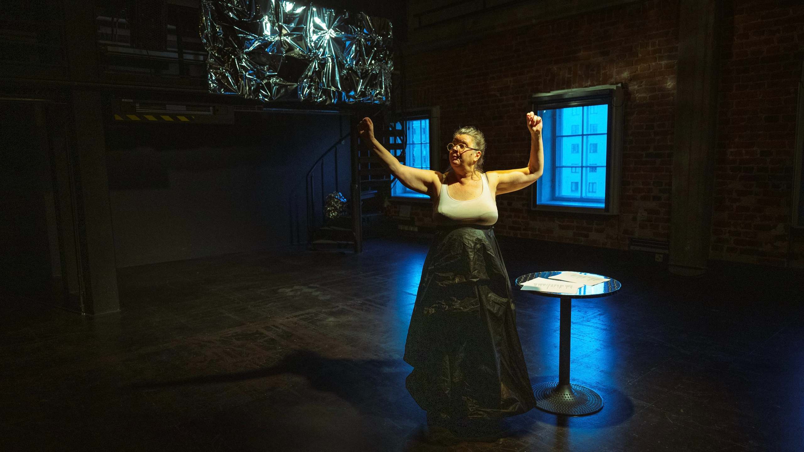 Frau in Bühnenkostüm mit erhobenem Arm steht vor leuchtenden blauen Fenstern im Theaterraum, ein Tisch mit Papier liegt im Vordergrund. Durch zwei Fenster schimmert blaues Licht herein.