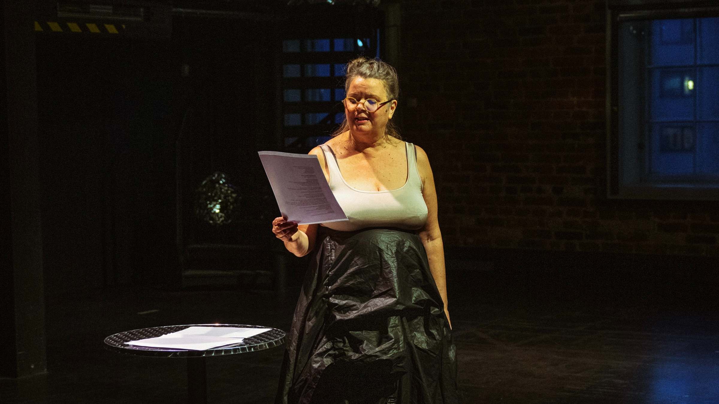 Eine Frau mit Zettel in der Hand blickt nach unten und steht an einem runden Tisch auf einer Theaterbühne mit Backsteinwänden.