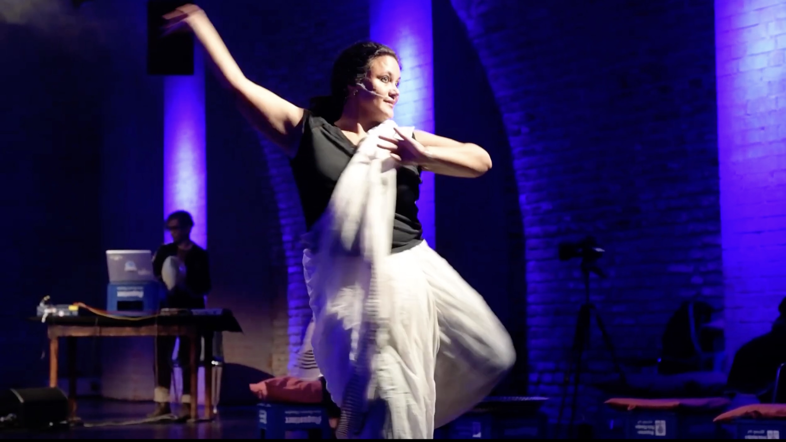 Eine Person in weißem, fließendem Kostüm tanzt auf einer Bühne und streckt einen Arm dynamisch nach oben, während der andere Arm seitlich gehalten ist; im Hintergrund sind blau‑violett angestrahlte Säulen, ein Tisch mit Technik und ein weiterer Mensch an einem Laptop zu sehen.