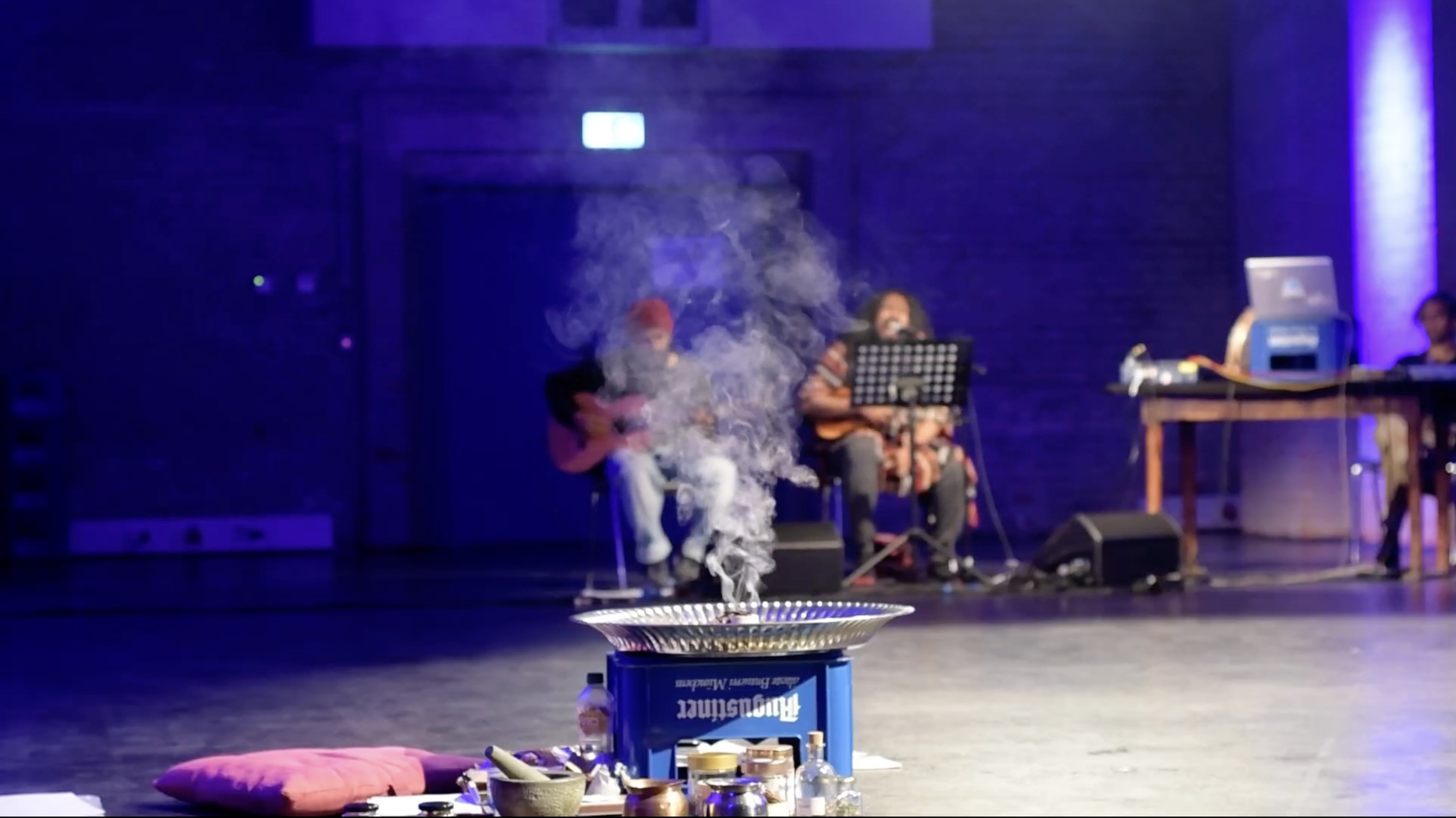 Im Vordergrund eines abgedunkelten Theaterraums steht auf dem Bühnenboden eine runde Metallfeuerschale auf einem blauen Gestell, aus der deutlich Rauch nach oben aufsteigt; davor liegen Kissen und verschiedene kleine Gefäße, im unscharfen Hintergrund sitzen mehrere Musikerinnen und Musiker mit Gitarren und Mikrofonen auf Stühlen, rechts ist ein Tisch mit Technik und Laptop zu sehen, die Bühne ist in blau violettes Licht getaucht.