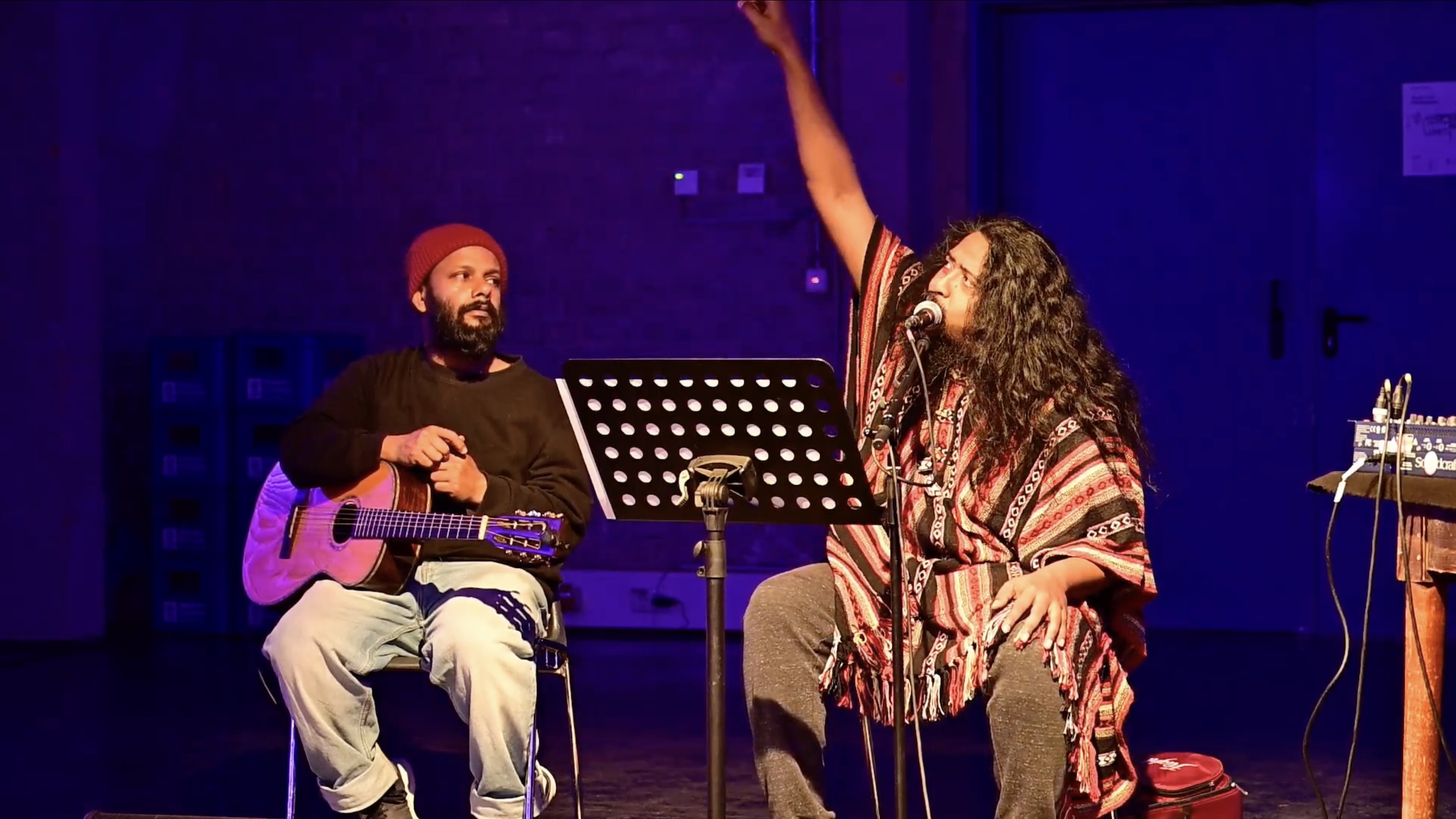 Zwei Musiker sitzen auf einer Bühne im blauen Licht; links spielt eine Person mit roter Mütze eine Gitarre, rechts singt eine Person mit langen Haaren ins Mikrofon und streckt einen Arm energisch nach oben, vor ihnen steht ein Notenständer.