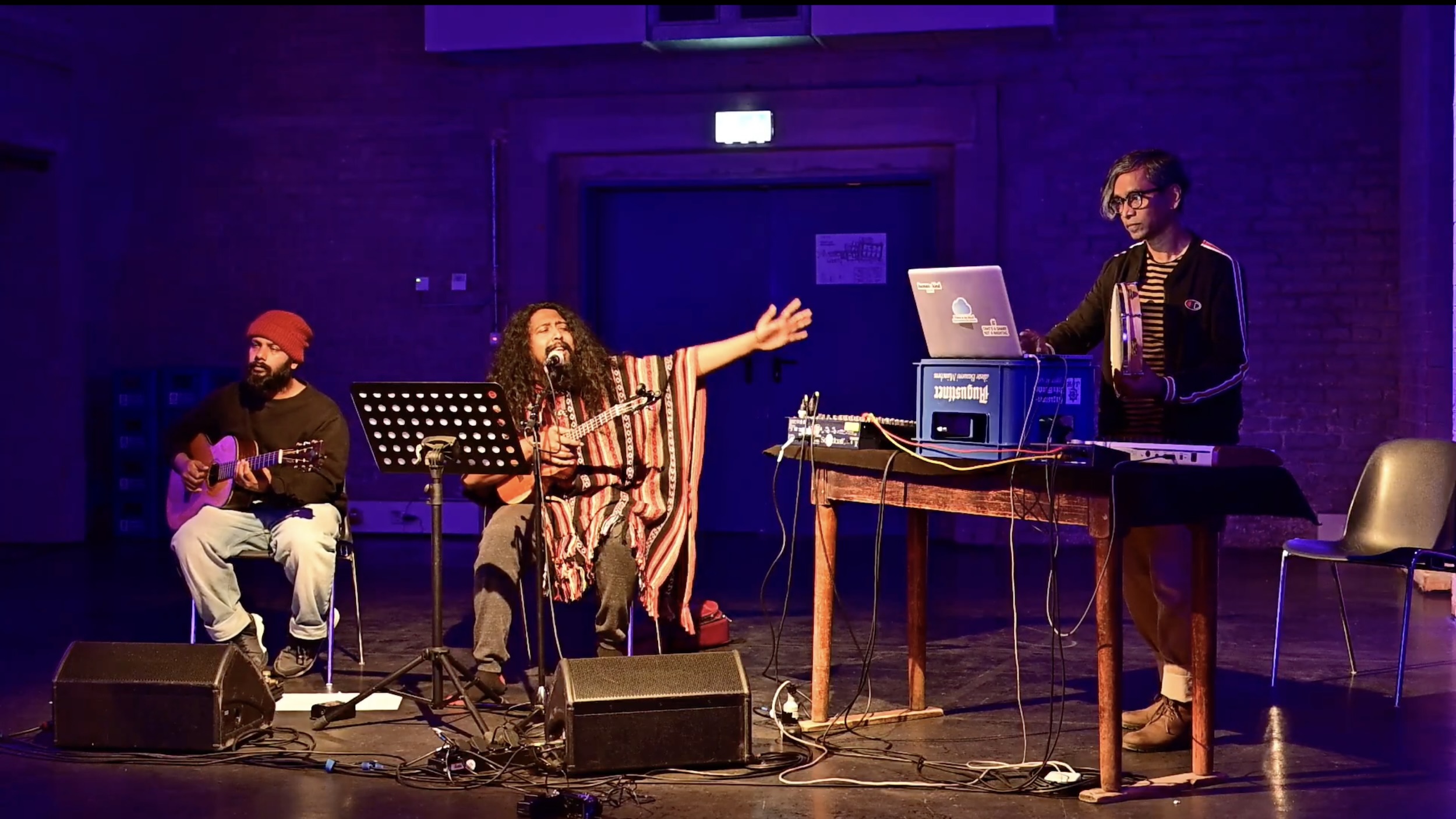 Drei Personen musizieren auf einer in blau‑violettes Licht getauchten Bühne: Links sitzt eine Person mit roter Mütze an der Gitarre, in der Mitte singt eine Person mit langen Haaren ins Mikrofon, rechts steht eine weitere Person hinter einem Tisch mit Laptop und Mischpult; vor den Musiker*innen liegen Kabel und Lautsprecher auf dem Boden.