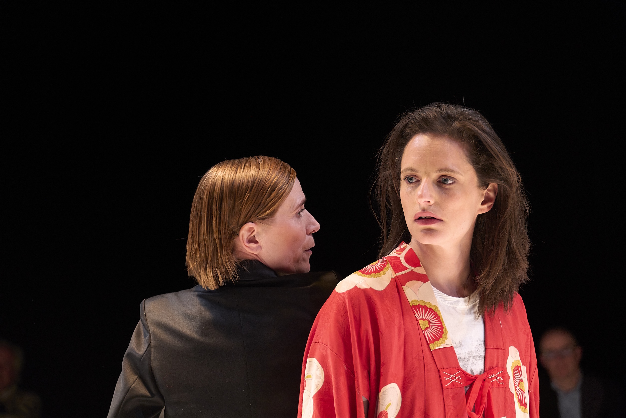 Zwei Personen stehen auf einer Bühne vor dunklem Hintergrund; eine Person im roten Oberteil blickt überrascht zur Seite, während die andere Person sich von hinten annähert und ihr etwas zuflüstert oder nahe spricht. Dramatische Theaterszene mit fokussierter Beleuchtung.