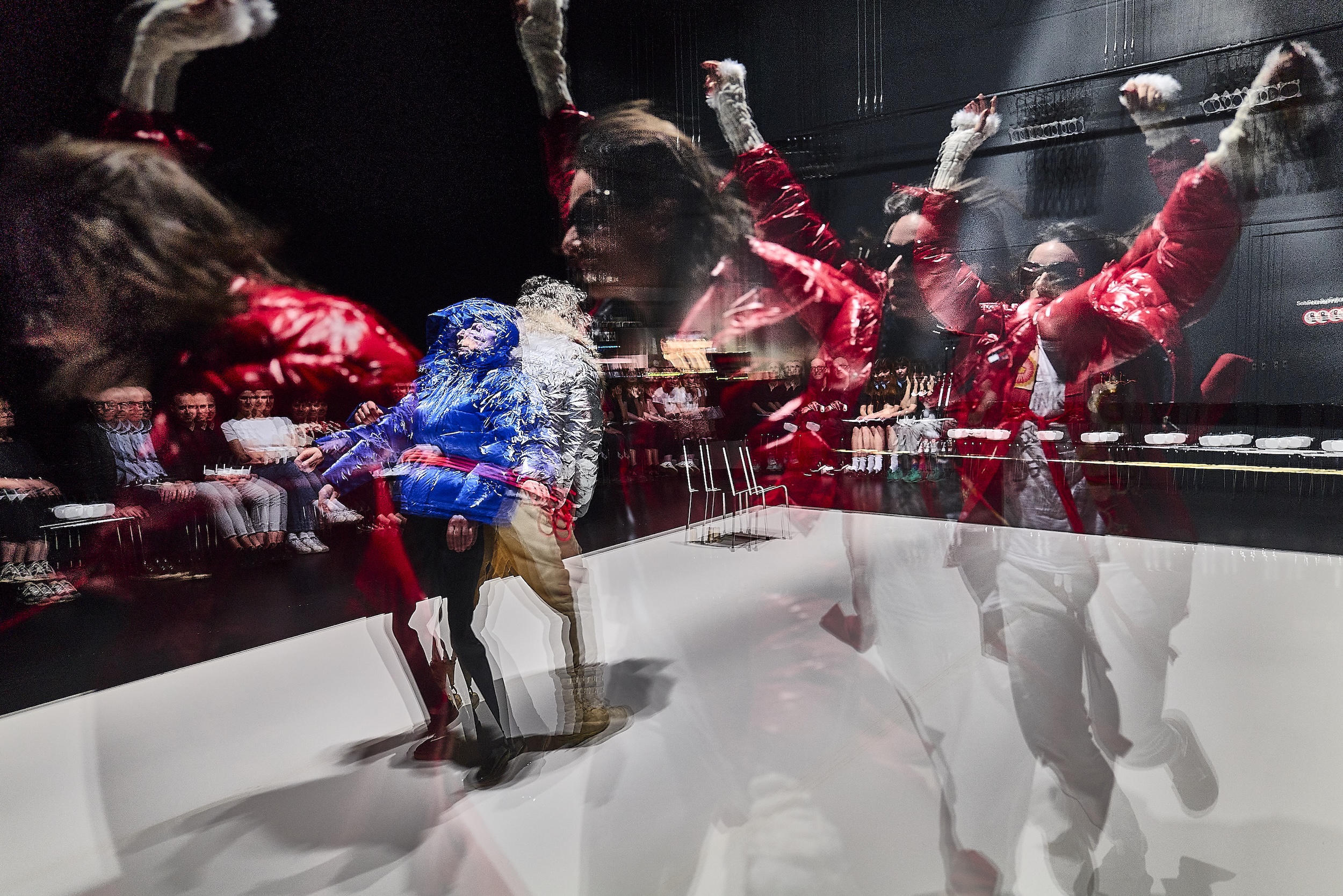 Person in blauem Outfit steht auf einer Bühne, umgeben von mehreren rot-schwarzen, tentakelartigen mechanischen Strukturen, die sich dynamisch um sie herum ausbreiten; futurale, performative Szene mit dramatischer Beleuchtung.