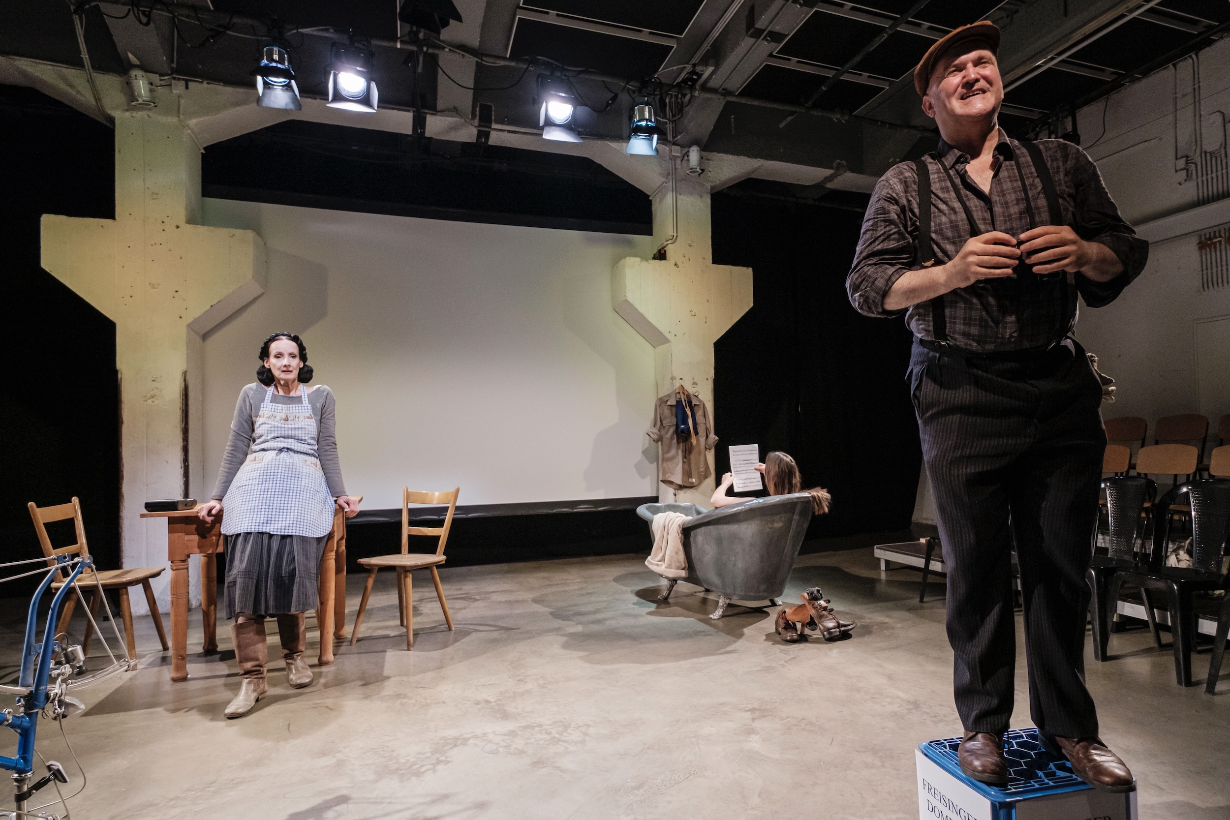 vorne rechts steht eine Person in Arbeitskleidung erhöht auf einer Kiste, links eine Person im Kleid und Schürze am Holztisch, im Hintergrund sitzt eine Person in einer Metallbadewanne und liest, darüber Scheinwerfer und Industriebühnenarchitektur.