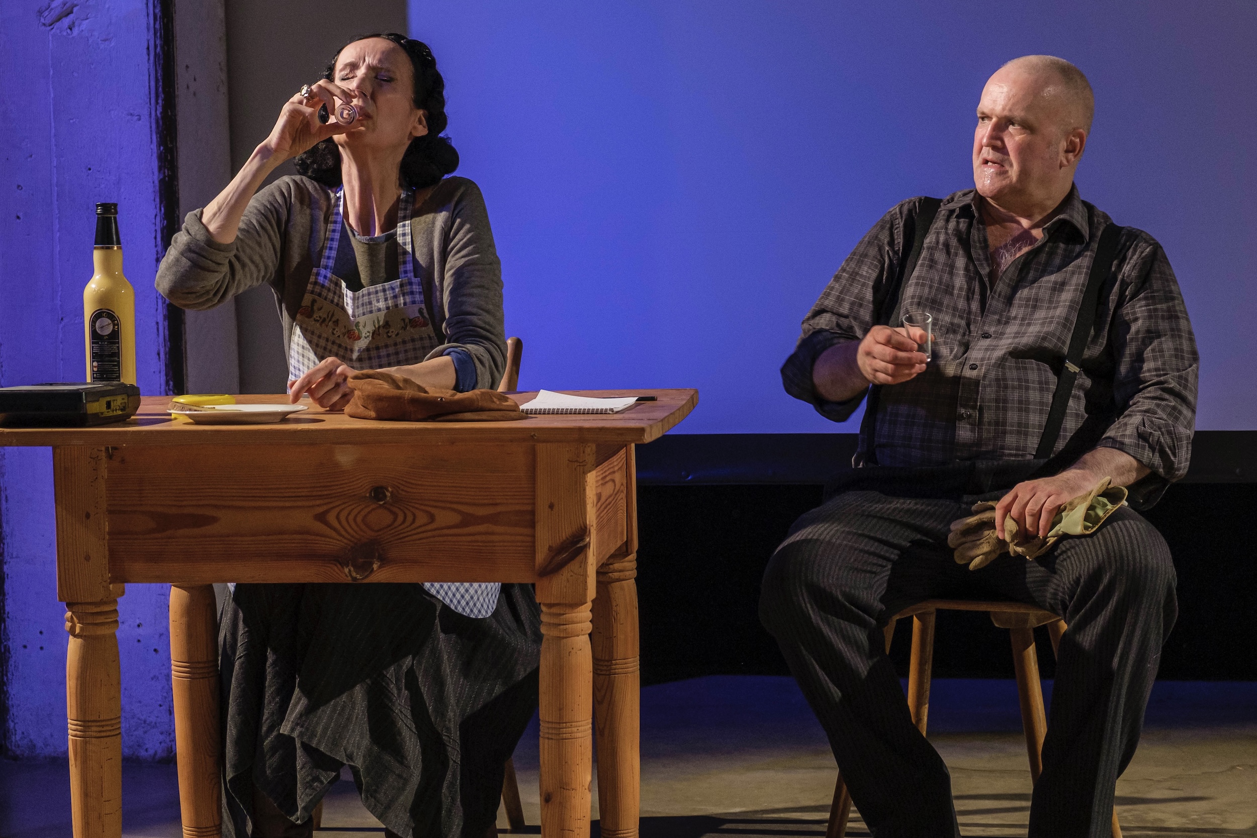 Zwei Personen sitzen an einem Holztisch in einer Theaterszene; links eine Person mit Schürze und Getränkeflasche, rechts eine Person in kariertem Hemd mit Hosenträgern, beide halten kleine Gläser und wirken im Gespräch vertieft.