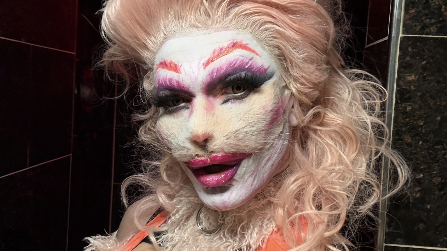 Eine Person mit voluminösem, blassrosa-lockigem Haar trägt ein flauschiges, helles Outfit mit orangem Halsband. Das Gesicht der Person weist Elemente von Fell wie bei einem Kaninchen auf. Der Hintergrund besteht aus dunklen Fliesen und gibt dem Bild eine leicht glamouröse Atmosphäre.