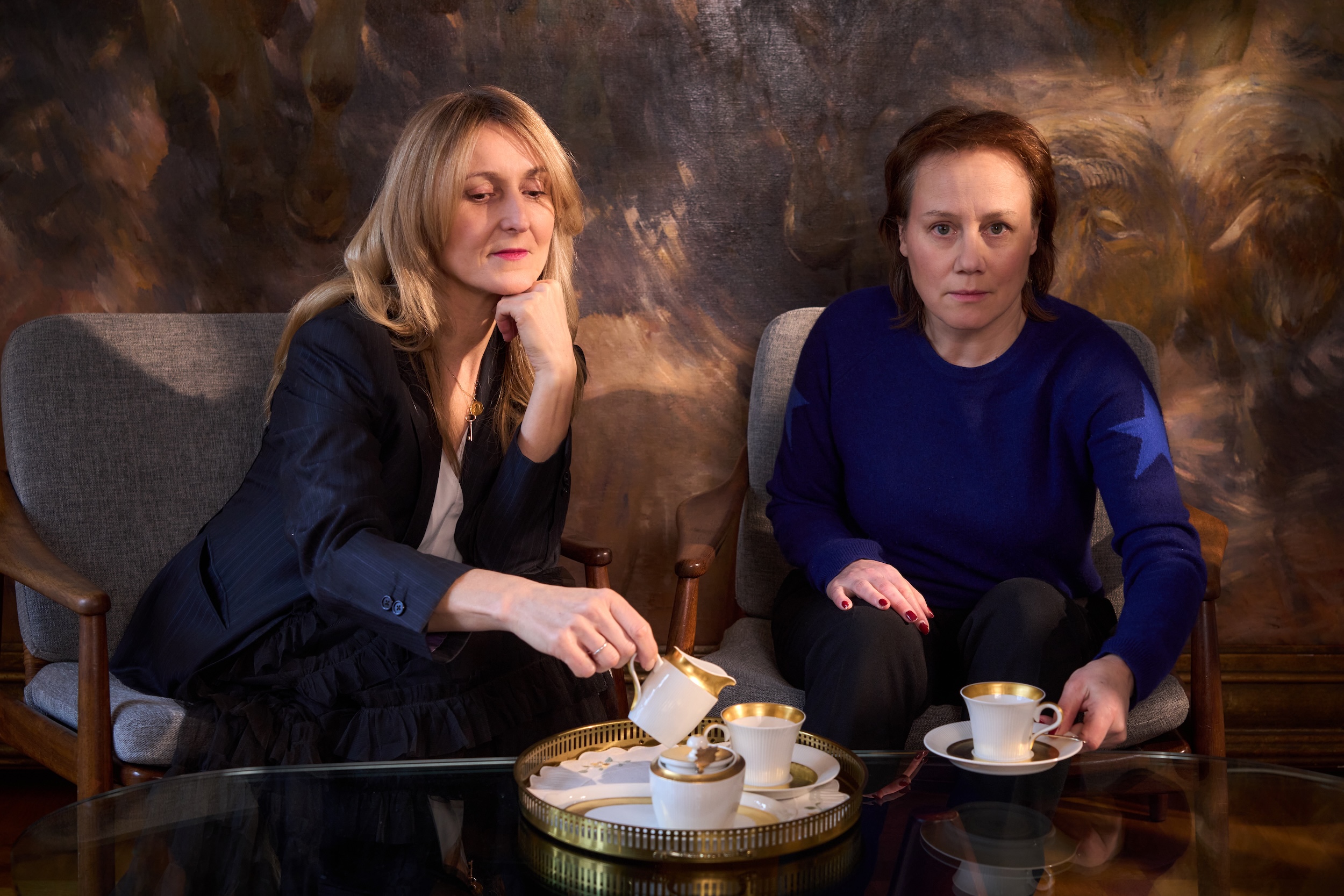 Zwei Personen sitzen einander zugewandt in einem Wohnzimmer‑Setting auf grauen Polstersesseln vor einem dunklen, malerischen Wandbild. Die Person links trägt einen dunklen Hosenanzug mit weißer Bluse und hält eine Kaffeekanne über ein weiß‑goldenes Service, das auf einem runden Tablett auf einem Glastisch steht. Die Person rechts trägt einen kobaltblauen Pullover mit Sternmotiven an den Ärmeln und eine dunkle Hose, sie hält eine weiße Kaffeetasse mit Untertasse in den Händen. Der Raum wirkt warm ausgeleuchtet, die Szene erinnert an ein intimes Gespräch im Salon oder an eine Teestunde in einem klassisch eingerichteten Wohnzimmer.