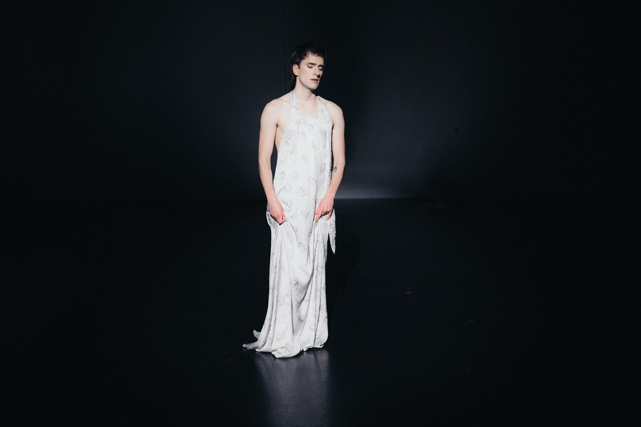 Eine schlanke Person im langen, hellen Kleid steht frontal und allein auf einer dunklen Theaterbühne, sie wird von einem Spotlicht aus dem schwarzen Raum herausgehoben.