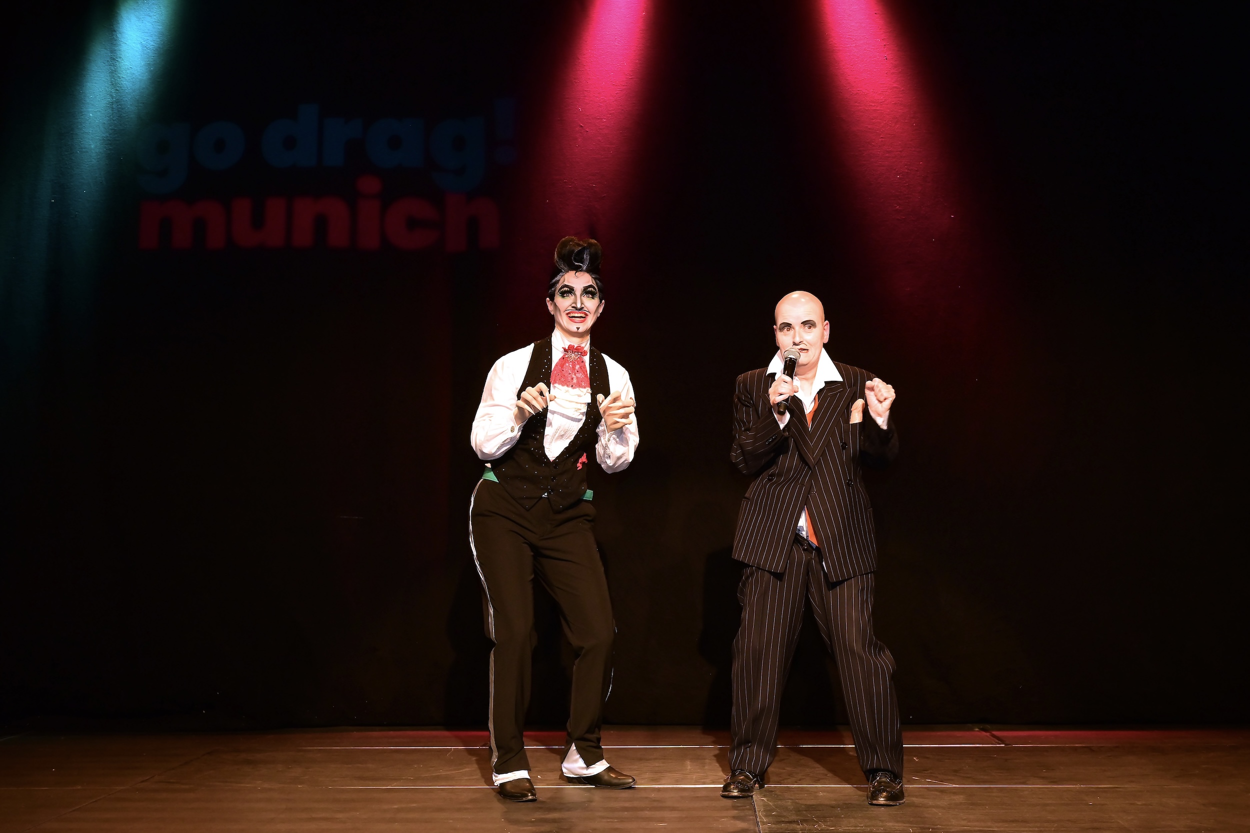 Zwei Drag-Performer in eleganten Anzügen stehen auf einer Bühne vor dunklem Hintergrund mit buntem Scheinwerferlicht und der unscharfen Aufschrift ‚go drag! munich‘, beide singen oder moderieren ins Mikrofon.
