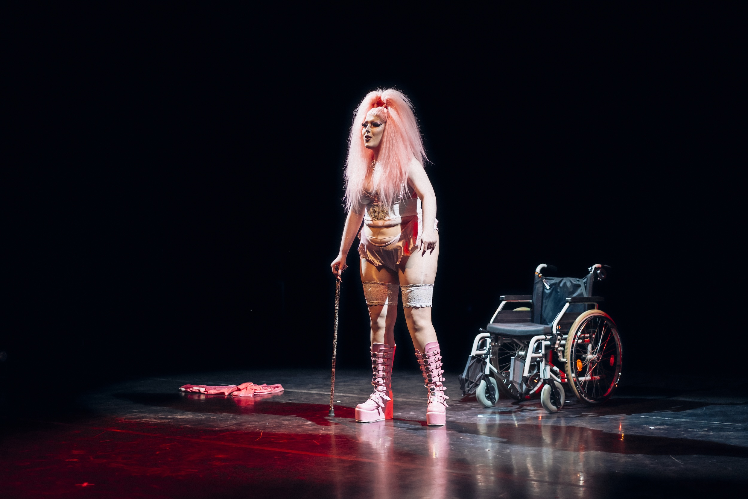 Drag-Performer*in mit pinker Perücke, Stützkorsett und Plateaustiefeln steht mit Gehstock im Lichtkegel auf einer dunklen Bühne, neben ihr ein leerer Rollstuhl und am Boden liegende Requisiten