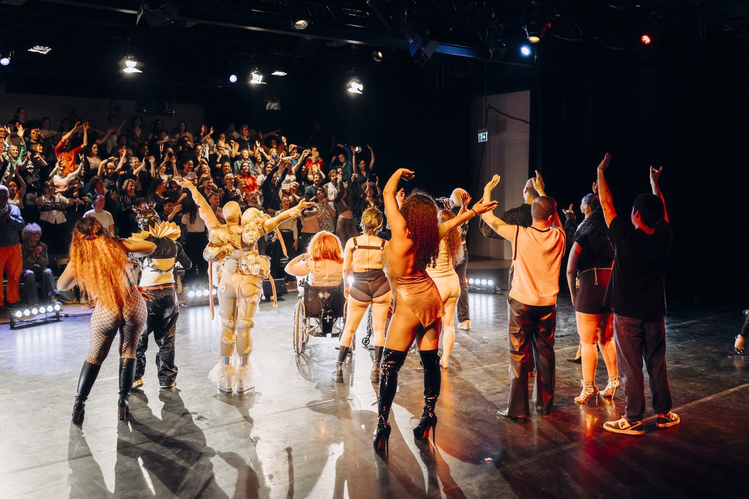 Zwei Personen stehen auf einer Bühne im Scheinwerferlicht, eine in dunklem Outfit mit kurzen hellen Haaren, die andere in einem futuristischen, gemusterten Kostüm mit Helm; beide agieren körperlich miteinander, als würden sie ein theatralisches oder comichaftes Drag-Duo spielen