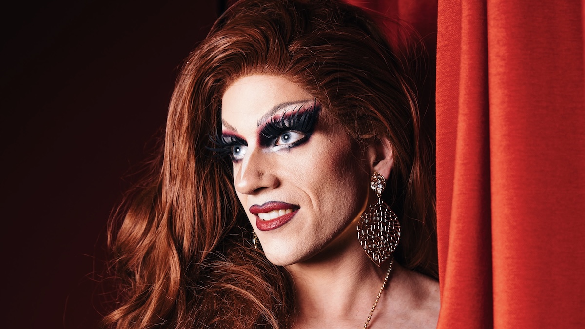 Eine Dragqueen steht vor einem roten Vorhang und trägt auffällige, goldfarbene, baumelnde Ohrringe sowie ein passendes Schmuckstück am Hals. Ihr langes, leicht gewelltes Haar ist rotbraun und fällt über die Schulter. Die Szene wirkt glamourös und erinnert an eine Theater- oder Bühnensituation.