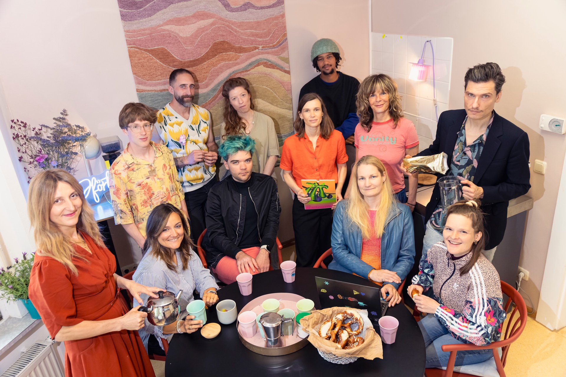 Gruppenfoto eines Teams in einem gemütlichen Innenraum mit warmem Licht und kreativer Atmosphäre. Mehrere Personen stehen und sitzen um einen runden Tisch mit Tassen, Gebäck und Getränken. Die Gruppe blickt überwiegend in die Kamera und trägt unterschiedlich farbenfrohe und individuelle Kleidung. Im Hintergrund sind Blumen, Kunstwerke und dekorative Lichtelemente zu sehen. Die Aufnahme wirkt lebendig, offen und gemeinschaftlich.