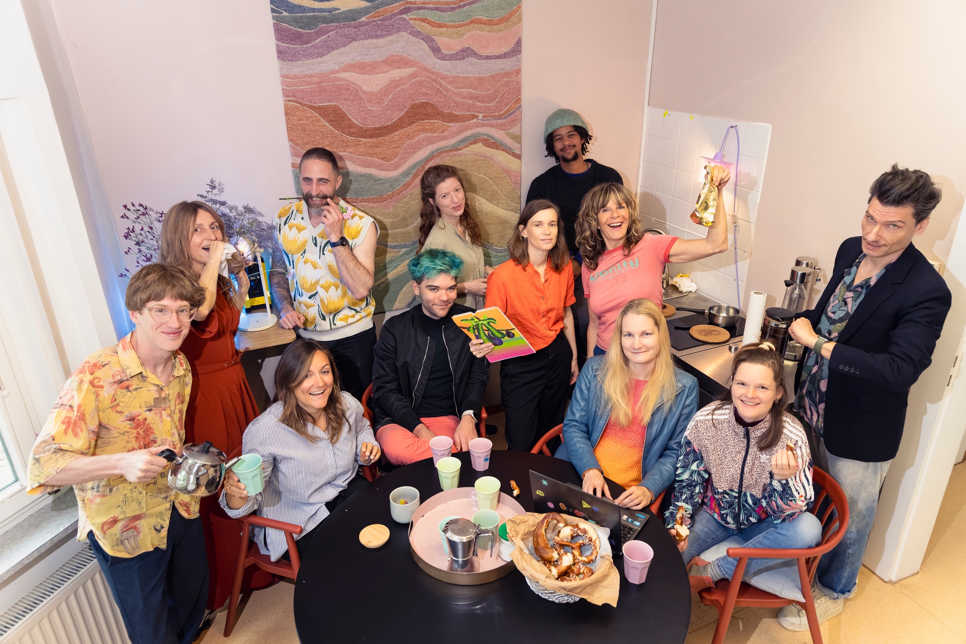 Lebendiges Gruppenfoto eines Teams in einer offenen Küche mit warmer und kreativer Atmosphäre. Mehrere Personen stehen und sitzen um einen runden Tisch mit Tassen, Brezeln und Getränken. Einige halten Gegenstände in der Hand oder lachen in die Kamera, wodurch die Szene spontan und dynamisch wirkt. Die Gruppe trägt farbenfrohe und individuelle Kleidung. Im Hintergrund sind ein großes abstraktes Kunstwerk, Blumen und dekorative Lichtquellen zu sehen. Die Aufnahme vermittelt Gemeinschaft, Kreativität und eine lockere Arbeitsatmosphäre.
