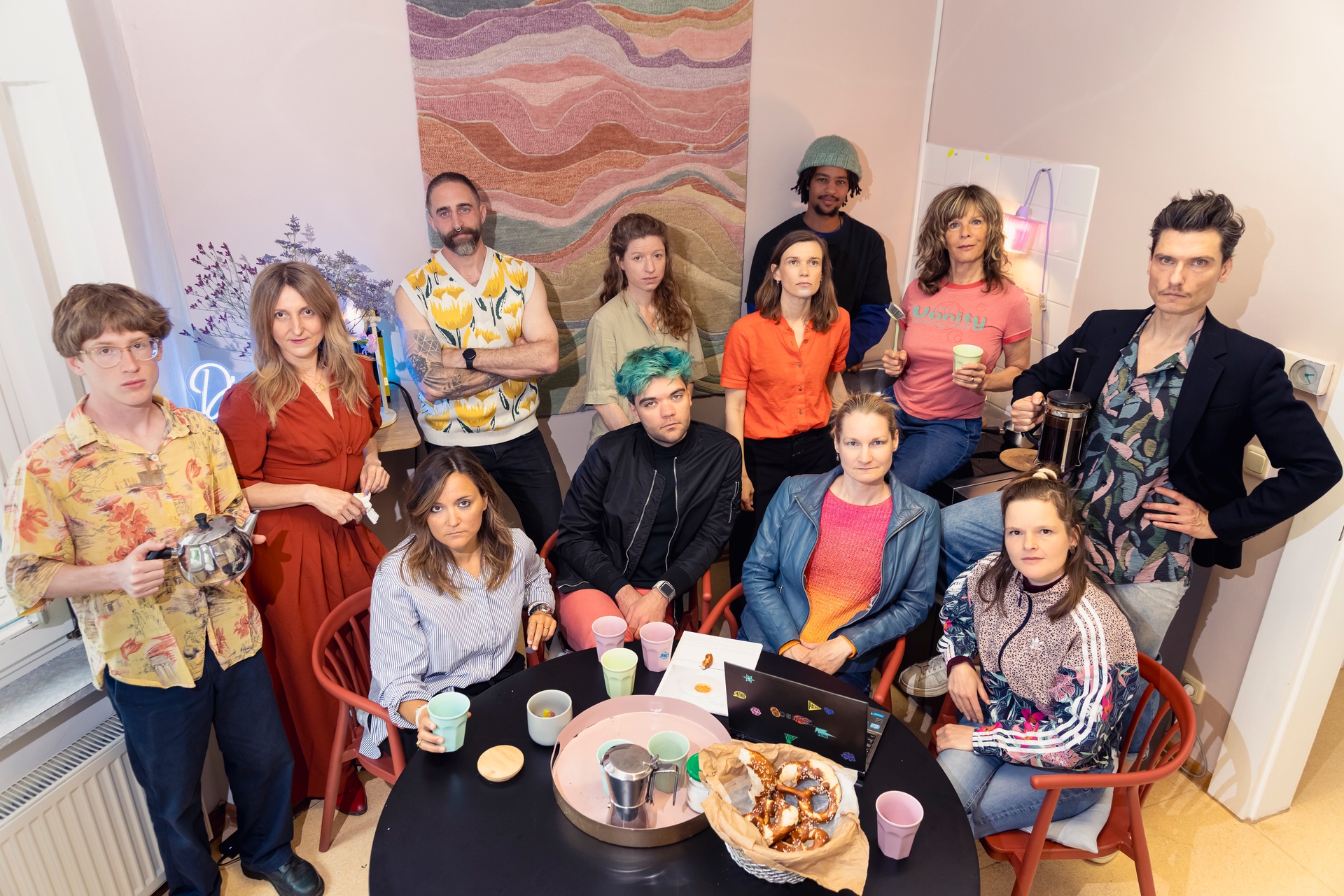 Gruppenfoto eines Teams in einem kreativ gestalteten Innenraum mit warmem Licht und gemütlicher Atmosphäre. Mehrere Personen stehen und sitzen um einen runden Tisch mit Tassen, Gebäck und einem Laptop. Die Gruppe blickt ernst oder neutral in die Kamera und trägt individuell farbenfrohe Kleidung. Im Hintergrund sind ein großes abstraktes Wandkunstwerk, Blumen und dekorative Lichtquellen zu sehen. Die Aufnahme wirkt modern, künstlerisch und gemeinschaftlich.