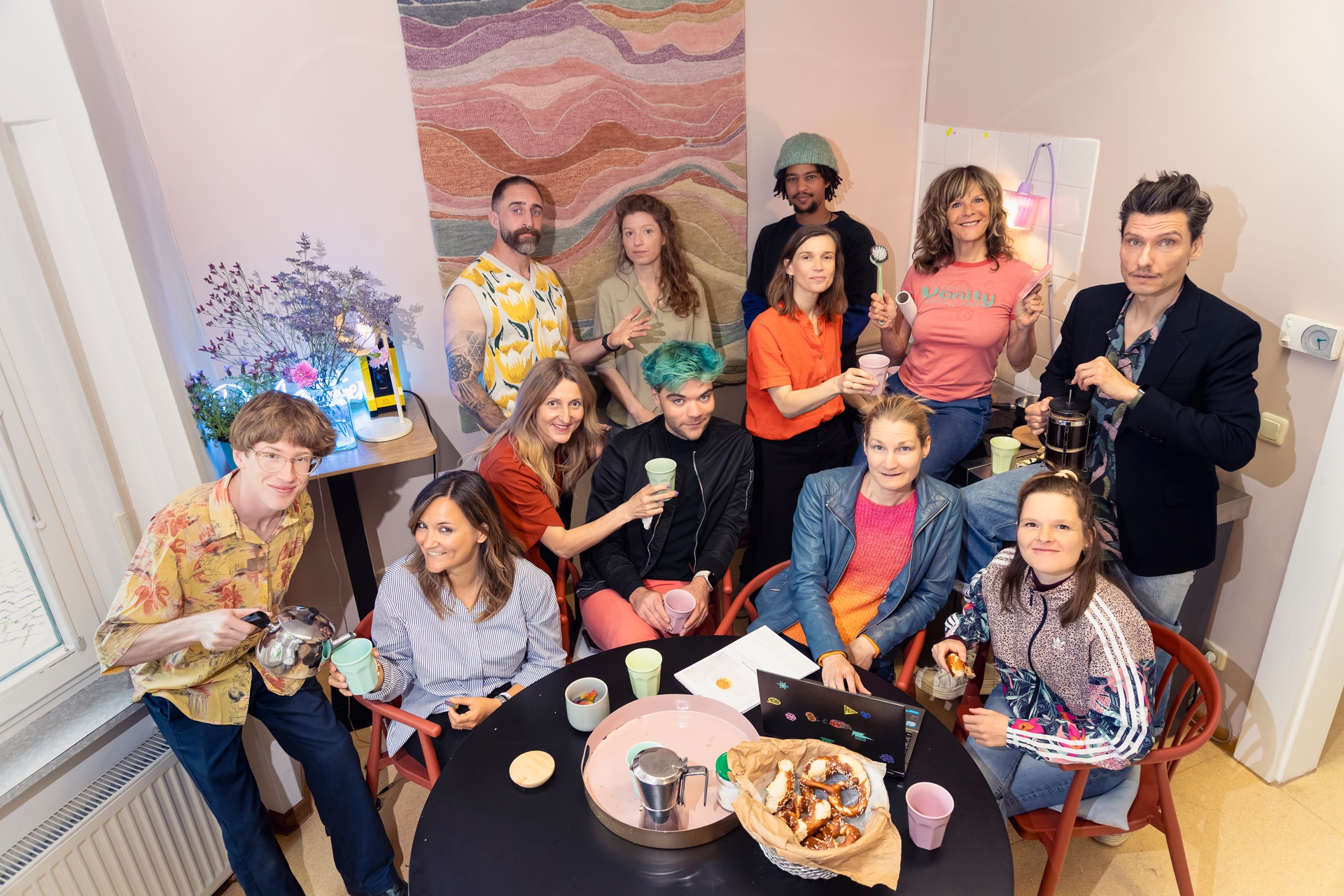 Lebendiges Gruppenfoto eines Teams in einem kreativ eingerichteten Innenraum mit warmer Beleuchtung. Mehrere Personen stehen und sitzen um einen runden Tisch mit Tassen, Brezeln, Getränken und einem Laptop. Einige halten Tassen oder Küchenutensilien in der Hand und lächeln in die Kamera, wodurch die Szene offen und spontan wirkt. Die Gruppe trägt farbenfrohe und individuelle Kleidung. Im Hintergrund sind Blumen, ein großes abstraktes Wandkunstwerk und dekorative Lichtquellen zu sehen. Die Aufnahme vermittelt Gemeinschaft, Kreativität und eine lockere, freundliche Atmosphäre.
