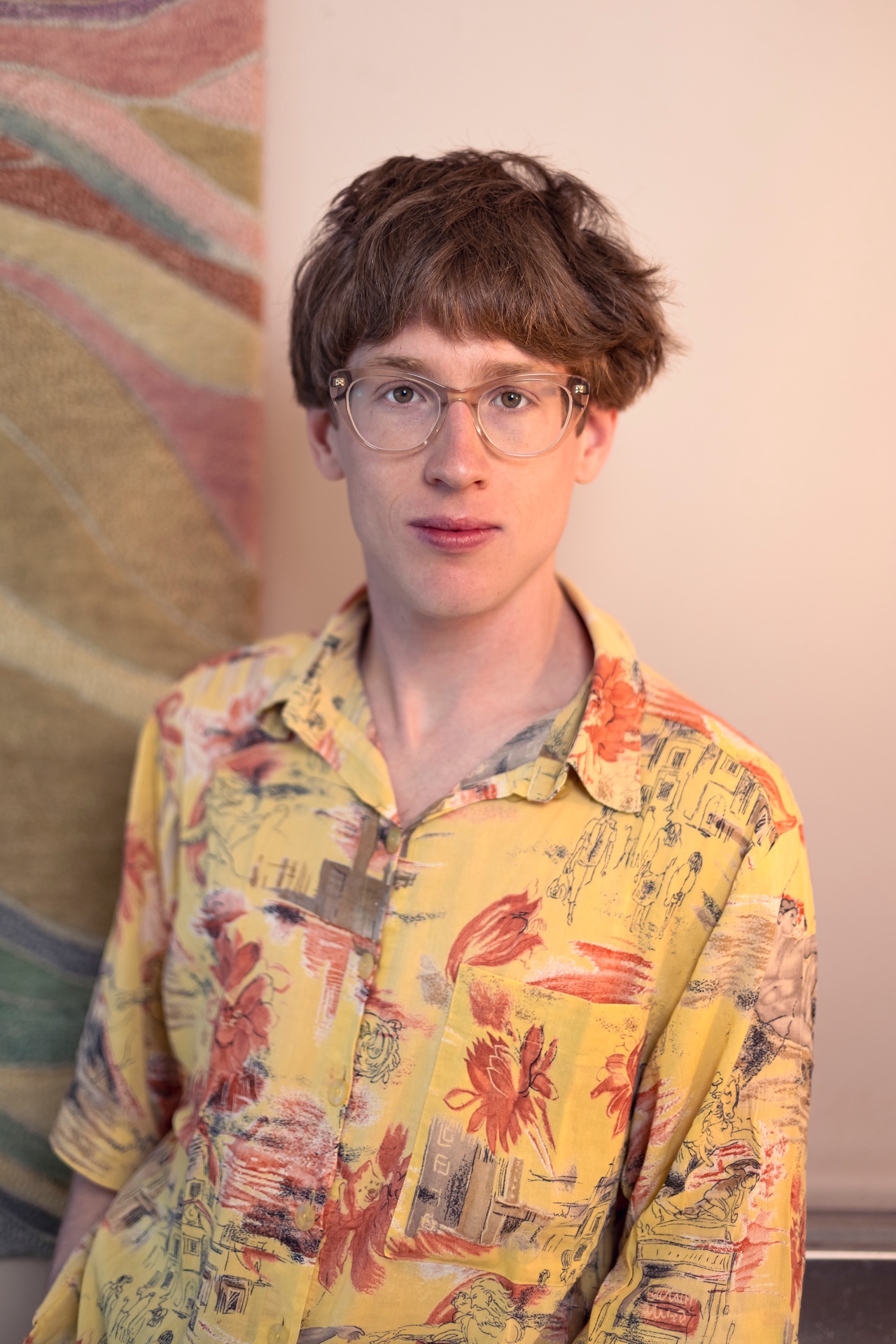 Eine Person mit kurzem, leicht zerzaustem braunem Haar und transparenter Brille blickt direkt in die Kamera. Sie trägt ein locker sitzendes, gelb gemustertes Kurzarmhemd mit floralen und illustrativen Motiven. Im Hintergrund sind eine pastellfarbene Wand und ein textiler Wandbehang mit wellenartigen Mustern zu sehen. Das Porträt wirkt ruhig, weich ausgeleuchtet und natürlich.