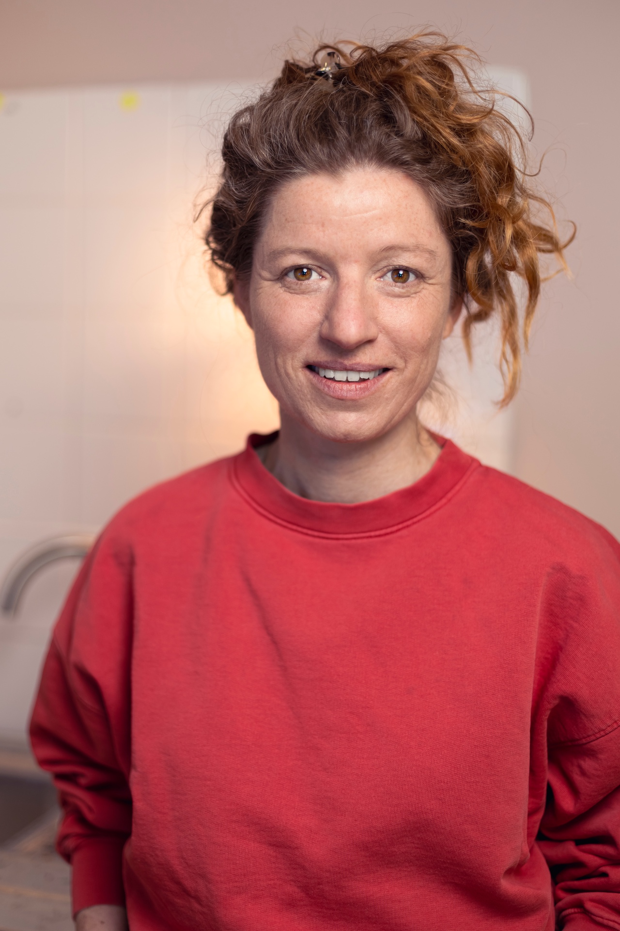 Eine Person mit lockigen rotbraunen Haaren und Sommersprossen steht lächelnd vor einem hellen, unscharfen Hintergrund. Sie trägt ein locker sitzendes rotes Sweatshirt, die Haare sind hochgesteckt, einzelne Locken fallen seitlich ins Gesicht. Das warme Licht und die geringe Tiefenschärfe verleihen dem Porträt eine freundliche, natürliche Atmosphäre.
