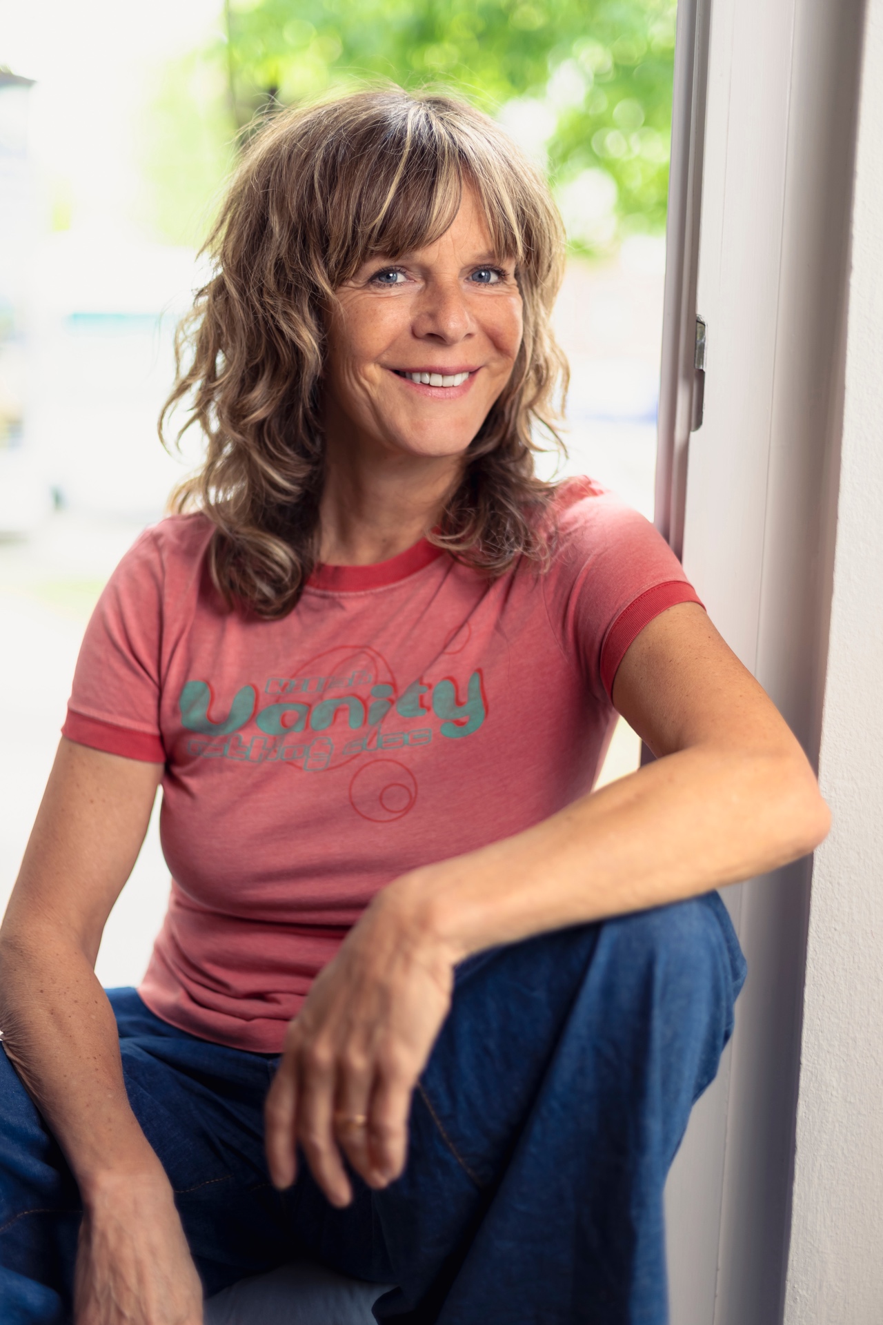 Porträtfoto einer lächelnden Frau mit schulterlangem, welligem Haar, die entspannt an einem Fenster sitzt und in die Kamera blickt. Sie trägt ein rosafarbenes T-Shirt mit Aufdruck und eine blaue Jeans. Durch das Fenster fällt natürliches Licht herein, im Hintergrund sind unscharfe grüne Pflanzen zu sehen. Die Aufnahme wirkt freundlich, lebendig und ungezwungen.