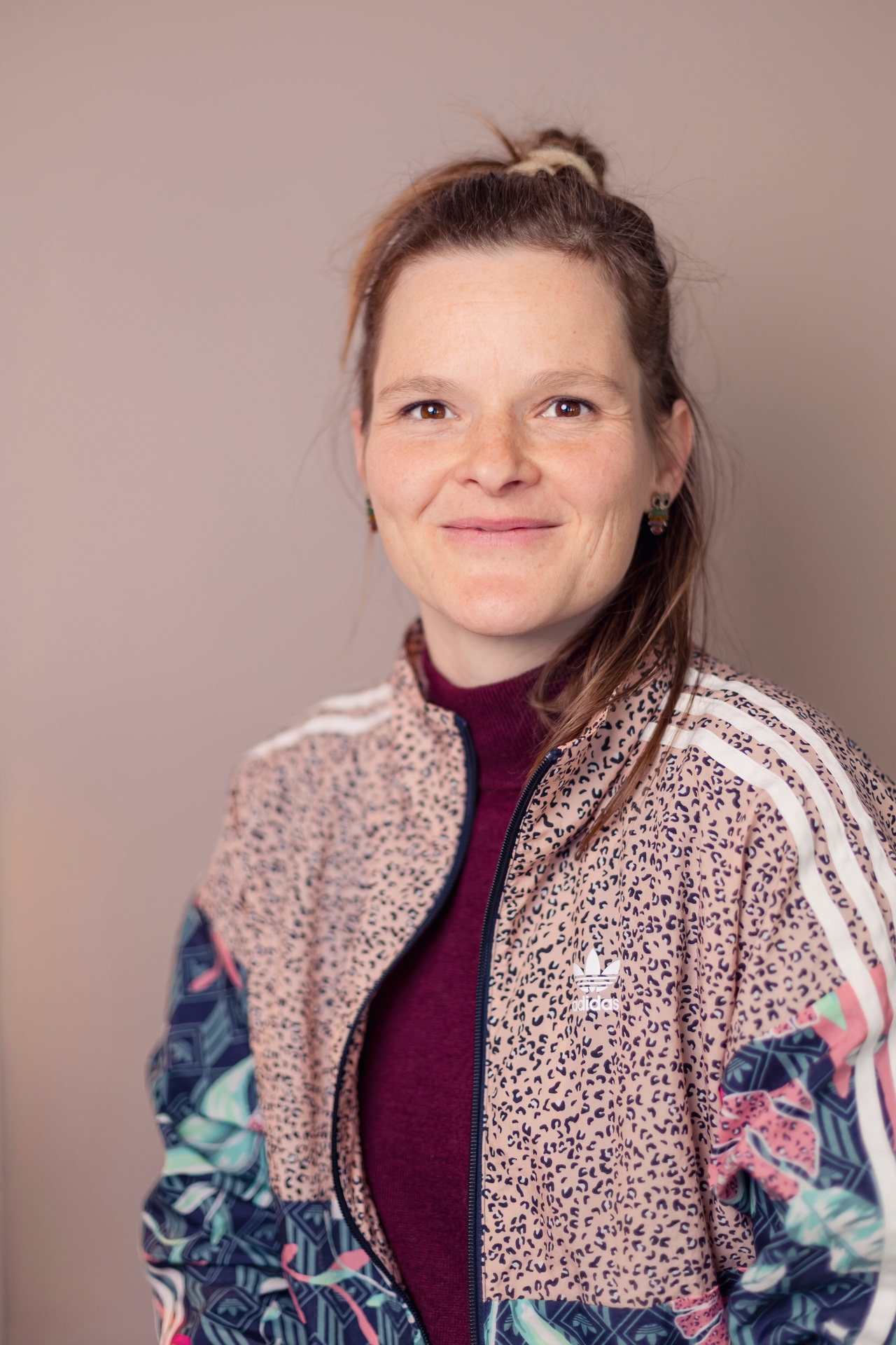 Porträtfoto einer lächelnden Person mit hochgebundenem Haar, die direkt in die Kamera blickt. Sie trägt eine gemusterte Sportjacke mit floralen und tierischen Mustern über einem dunkelroten Oberteil. Der schlichte, weich beleuchtete Hintergrund in warmen Farbtönen lenkt den Fokus auf den freundlichen Gesichtsausdruck. Die Aufnahme wirkt entspannt, modern und sympathisch.