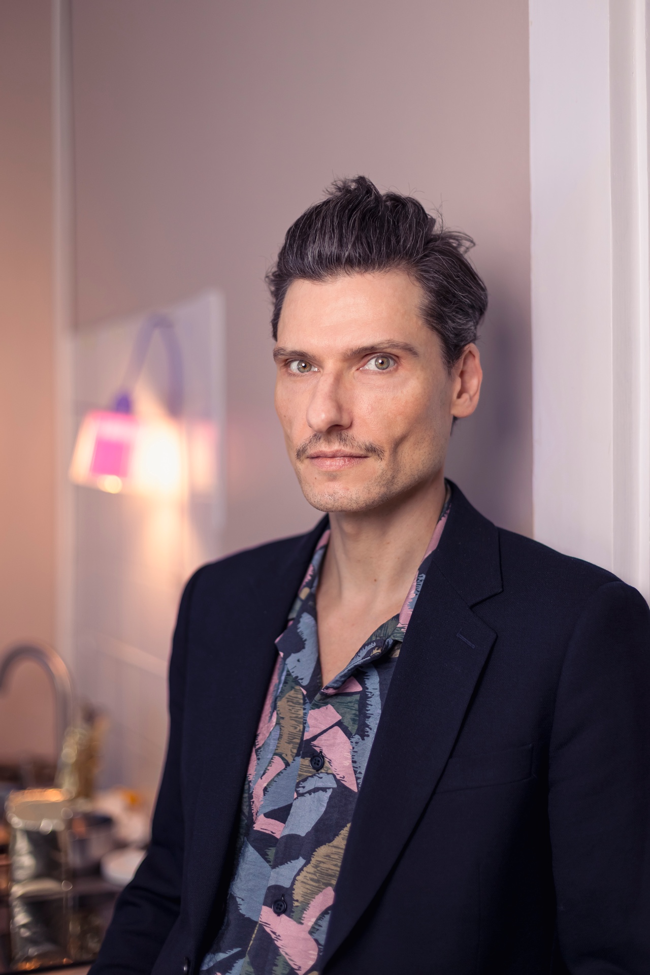 Porträtfoto einer Person mit kurzen dunklen Haaren und schmalem Schnurrbart, die direkt in die Kamera blickt. Die Person trägt ein dunkles Sakko über einem gemusterten Hemd in gedeckten Farben. Im Hintergrund sind unscharfe Innenraumelemente und warmes Licht zu sehen, was dem Bild eine stilvolle und moderne Atmosphäre verleiht.