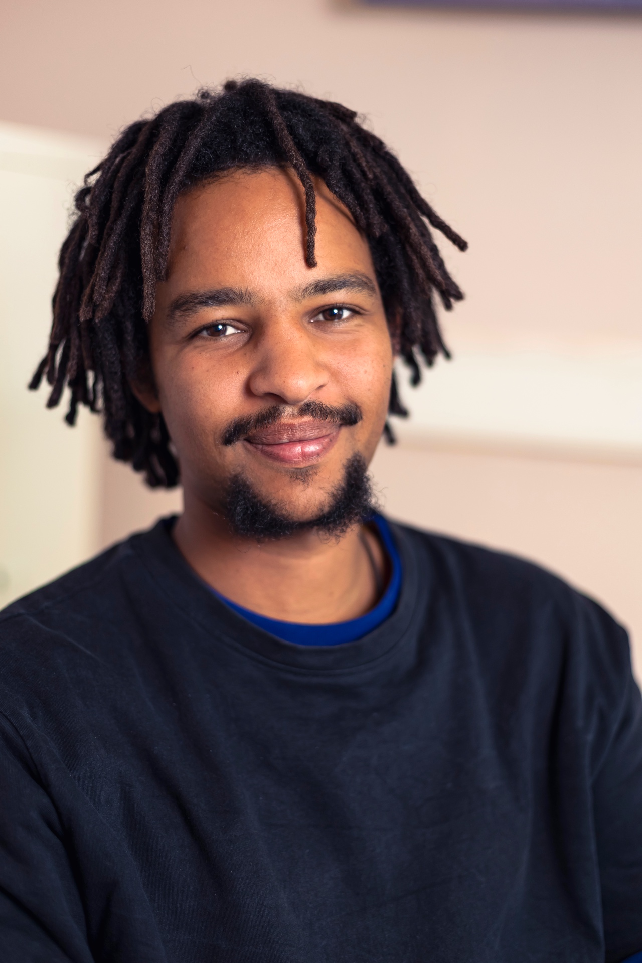 Porträtfoto eines lächelnden Mannes mit mittellangen Dreadlocks und kurzem Bart, der direkt in die Kamera blickt. Er trägt ein dunkles Sweatshirt über einem blauen Oberteil. Der helle, weich verschwommene Hintergrund lenkt den Fokus auf den freundlichen Gesichtsausdruck. Die Aufnahme wirkt ruhig, offen und natürlich.