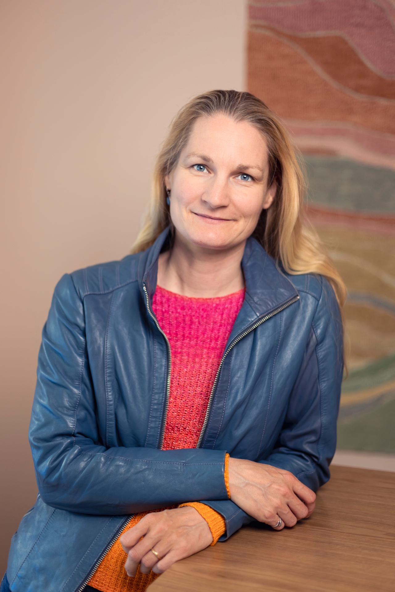 Porträtfoto einer Frau mit langen blonden Haaren und blauen Augen, die leicht lächelnd in die Kamera blickt. Sie trägt eine blaue Lederjacke über einem pink-orangen Strickpullover und lehnt entspannt an einem Holztisch. Im Hintergrund ist ein weich verschwommenes abstraktes Gemälde in warmen Farben zu sehen. Die Aufnahme wirkt freundlich, modern und professionell.
