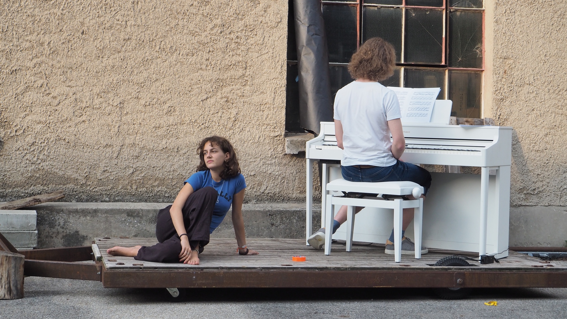  Zwei Performer – eine Person sitzt auf einem Holzwagen, die andere spielt Klavier im Freien vor einer alten Wand mit Fenster.