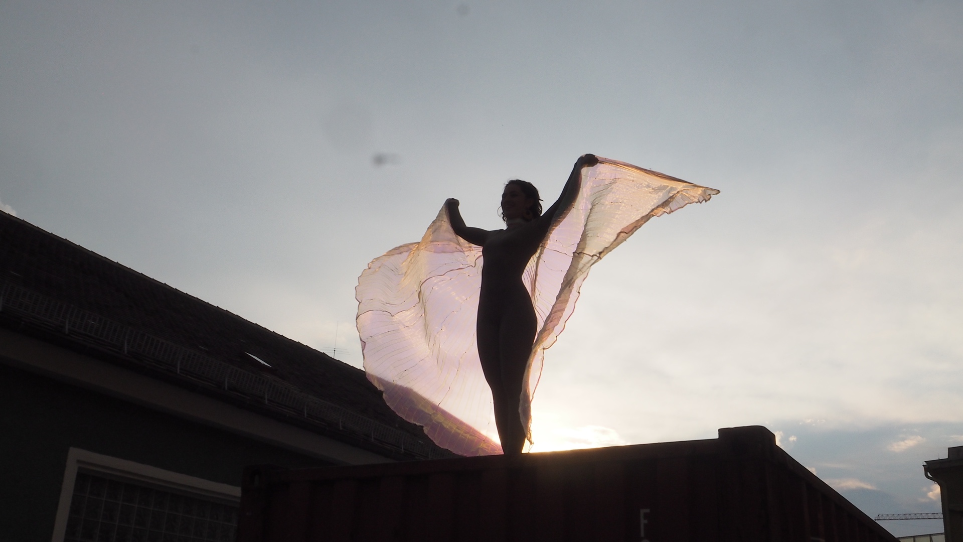 Silhouette einer Person mit ausgebreiteten, transparenten Tüchern, drapiert als Flügel, auf einem Containerdach vor Abendhimmel.